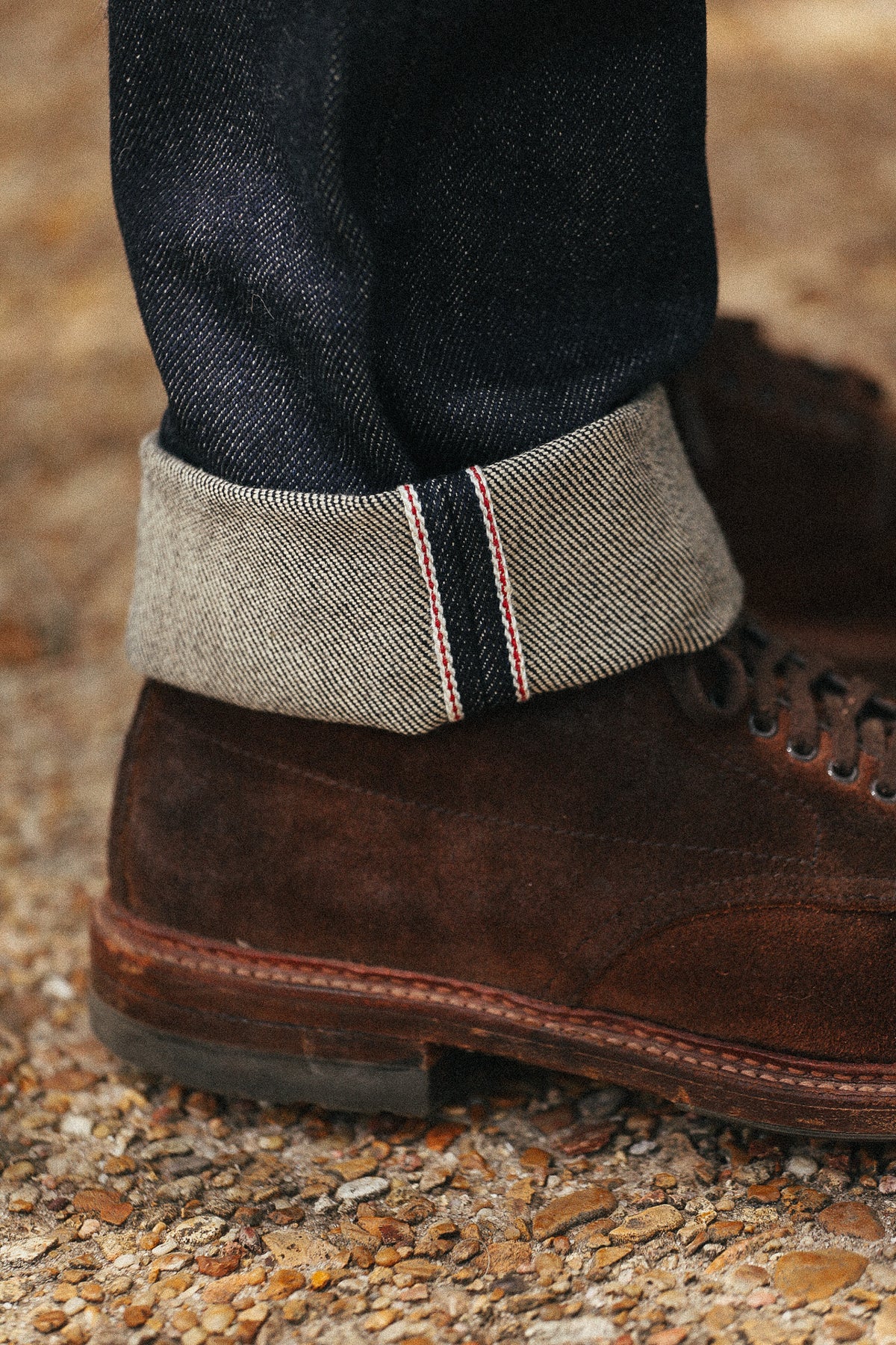Close-up of a person's ankle wearing a rolled-up pair of dark blue jeans with a visible herringbone twill cuff, paired with a brown suede lace-up boot on a gravel surface.