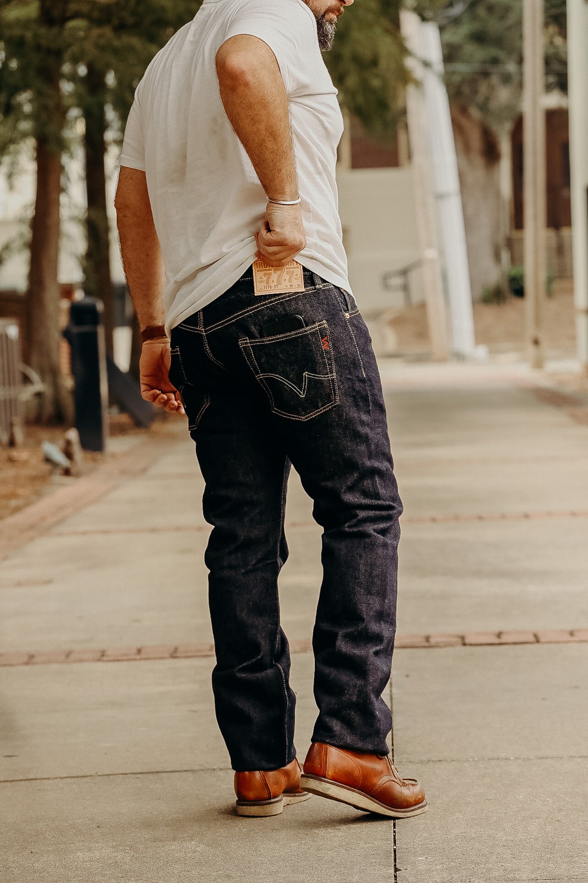 A person standing on a sidewalk wearing a white t-shirt and the 21 oz Selvedge Denim Super Slim Tapered Jeans in indigo color with brown shoes.