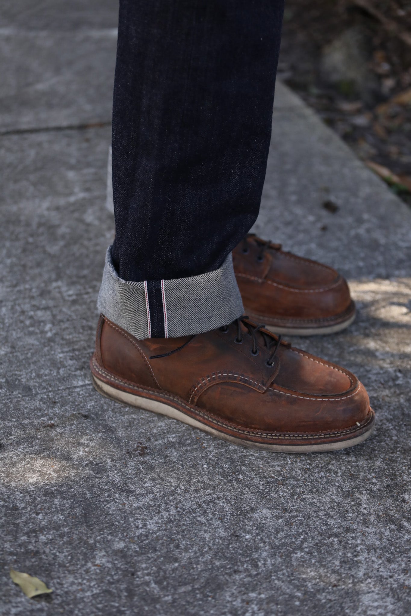 Brown leather moc toe shoes paired with medium indigo selvedge denim jeans cuffed on concrete pavement