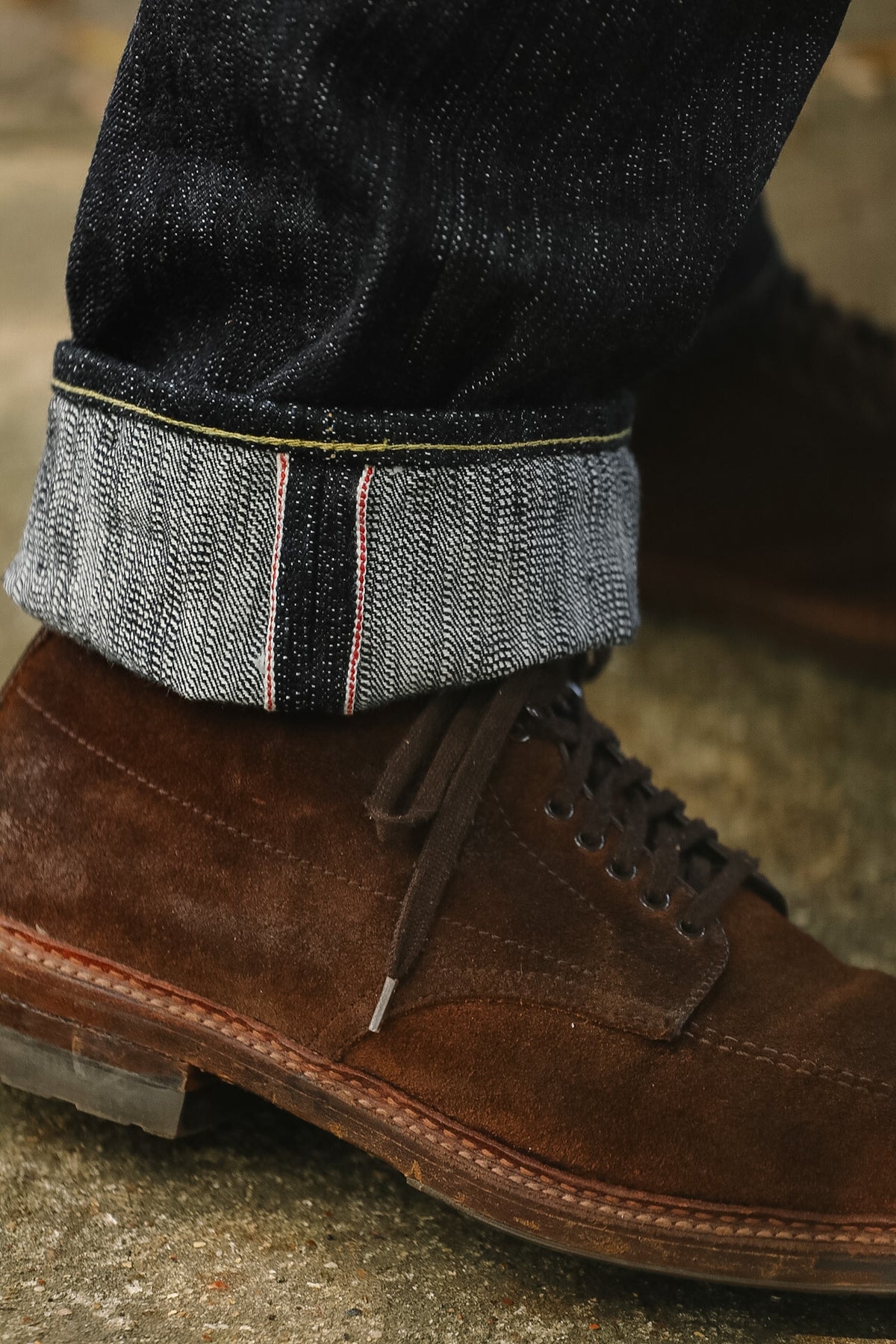 Close-up of a person's foot wearing a brown suede lace-up shoe and dark indigo jeans with a rolled cuff showing a gray and black herringbone pattern.