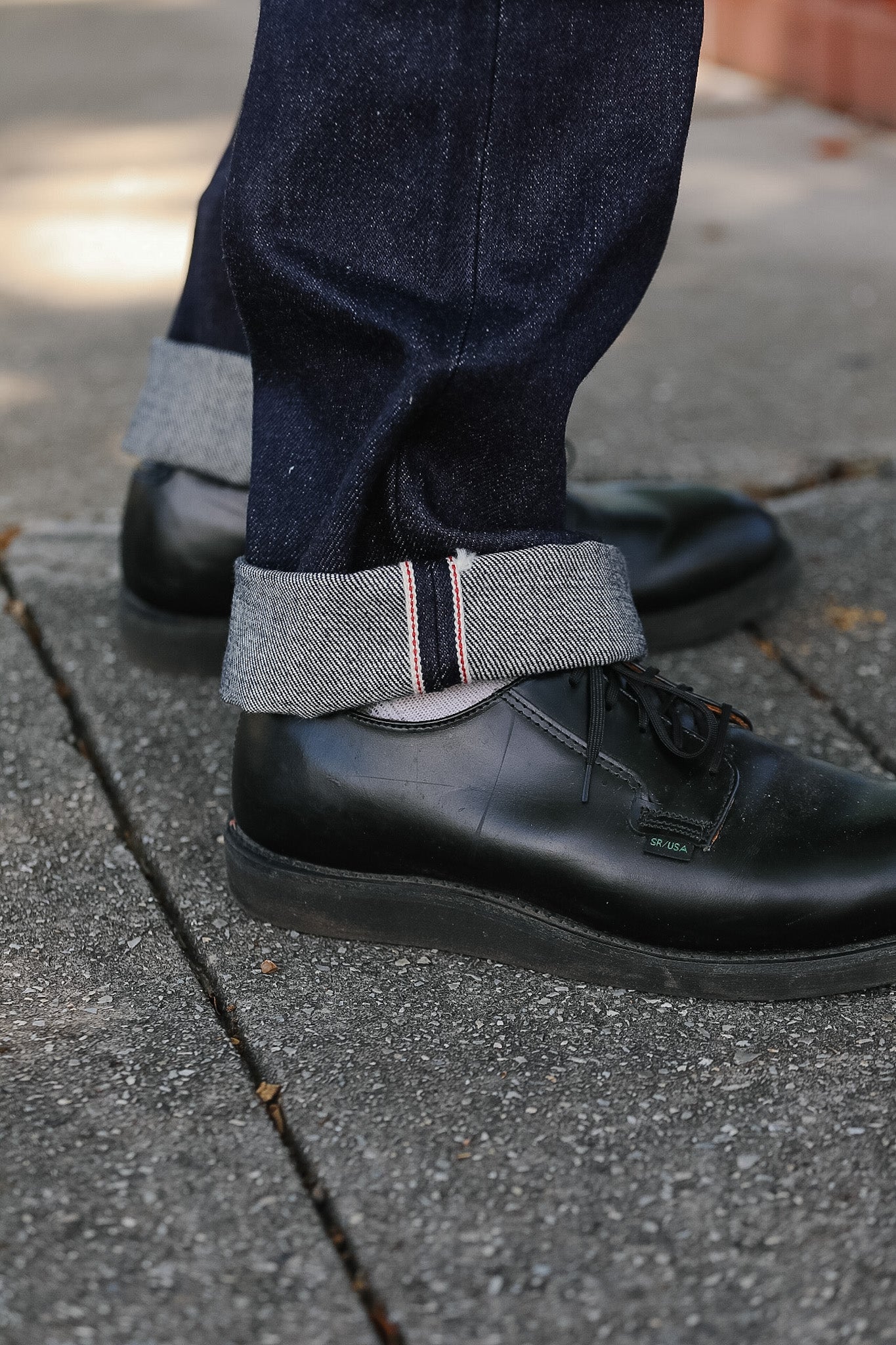 Close-up of black leather shoes and navy pants with rolled-up cuffs on a sidewalk.