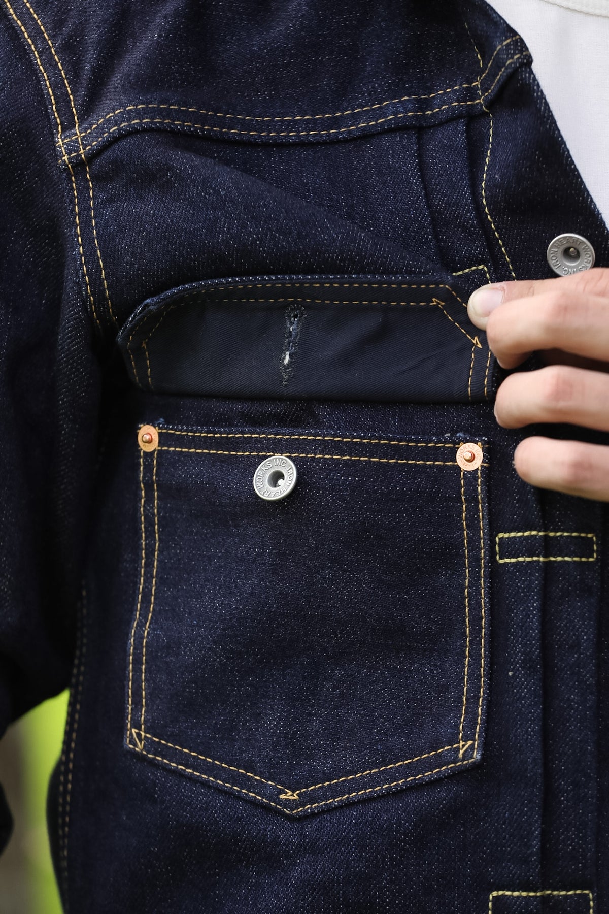 Close-up of a dark indigo denim jacket with a front pocket, contrast yellow stitching, and a hand holding the button closure.
