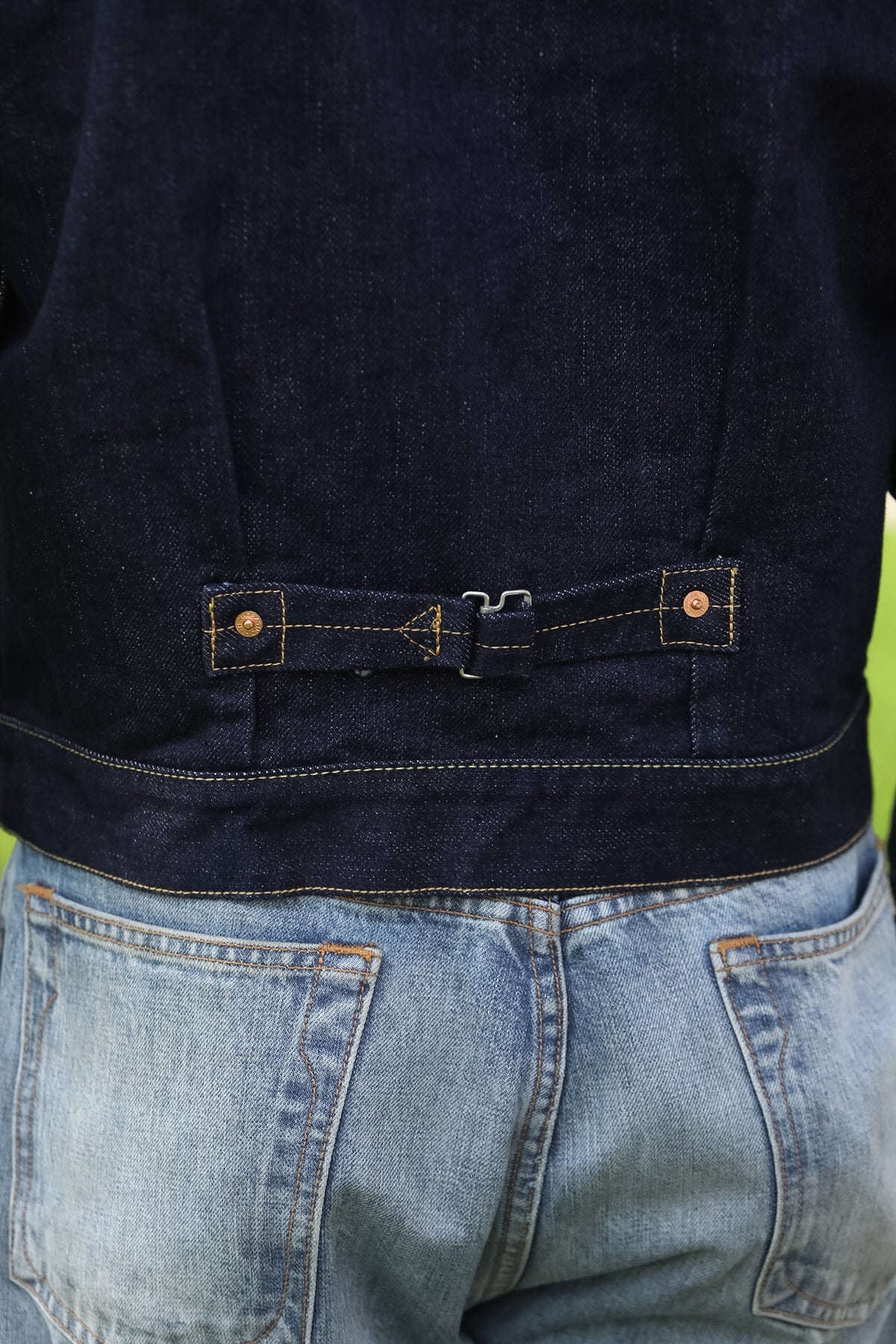 Close-up of the back of a dark indigo denim jacket worn over light blue jeans, showing a rear belt detail with metal buckle and contrast stitching.