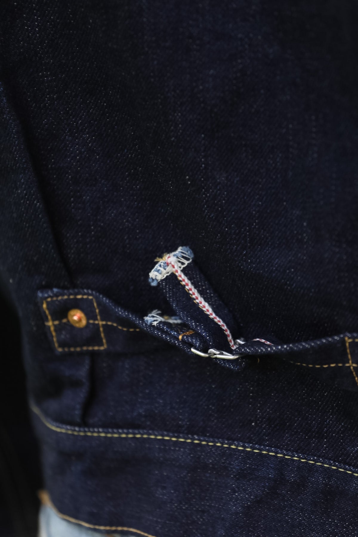 Close-up detail of dark indigo denim fabric showing a buttoned pocket with contrast yellow stitching and a decorative metal pull tab.