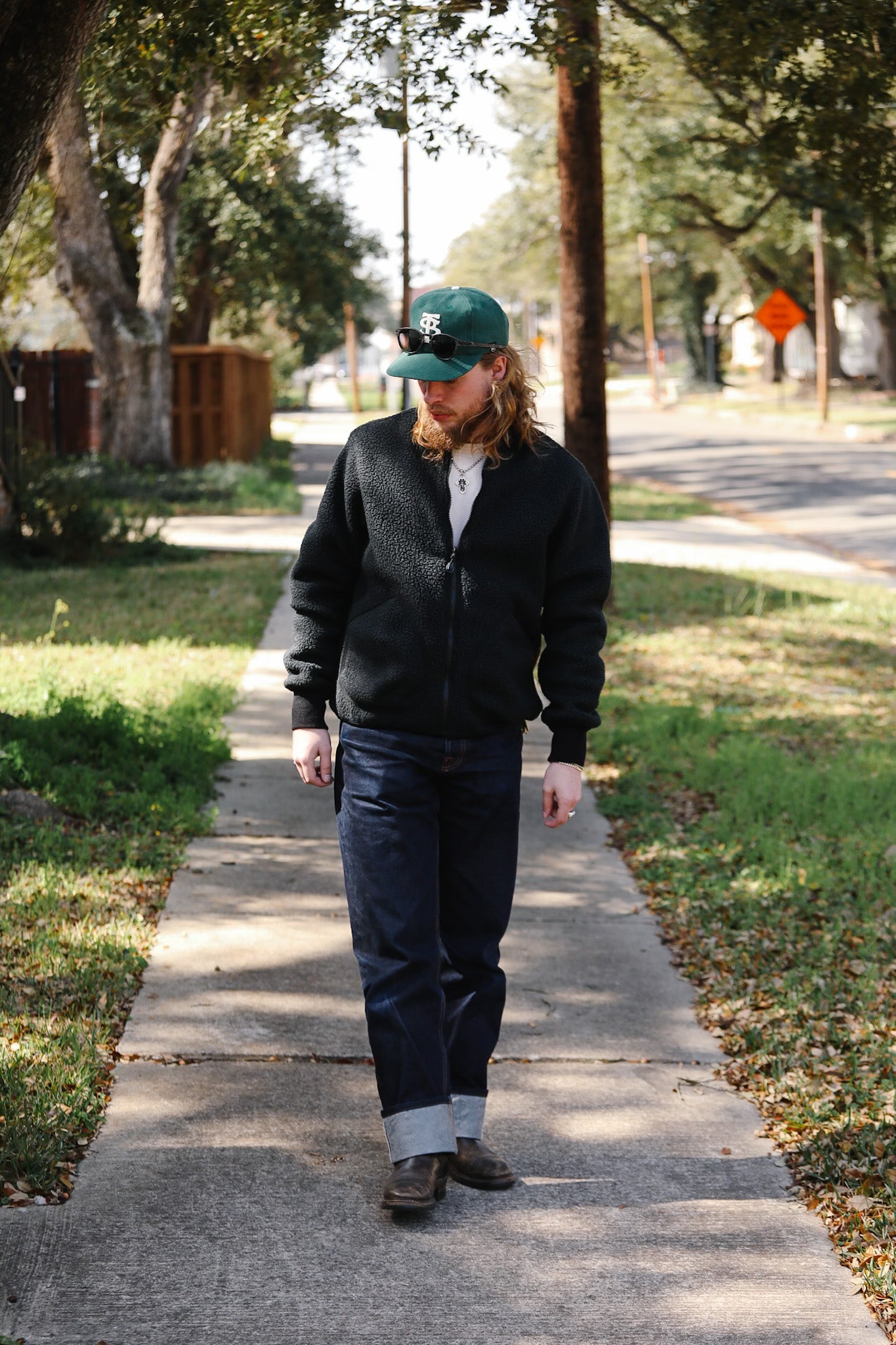 Man walking on a sidewalk wearing a black jacket, green cap, and blue jeans.