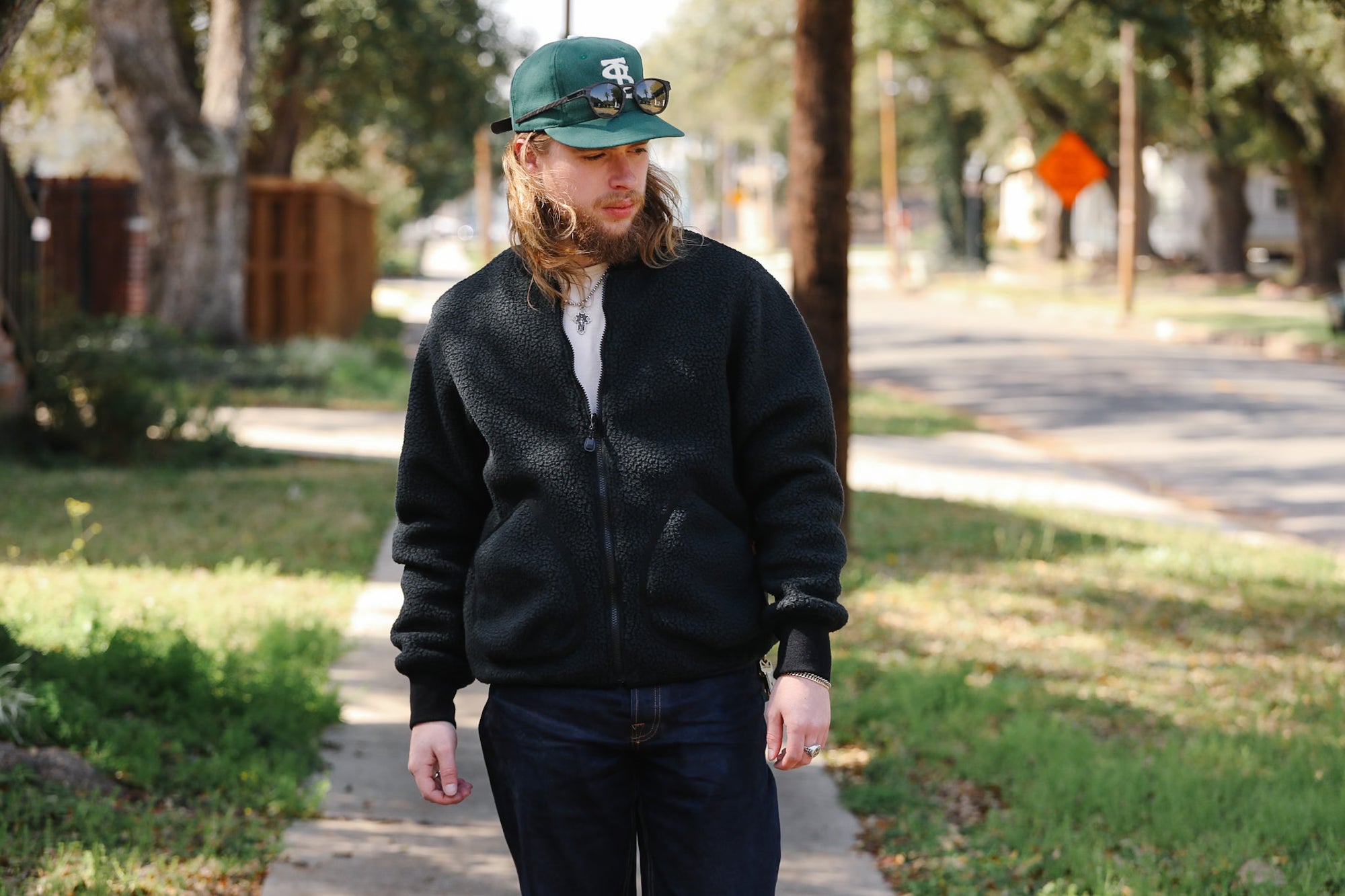 Person wearing a black jacket and green cap walking on a sidewalk with trees and a road in the background.