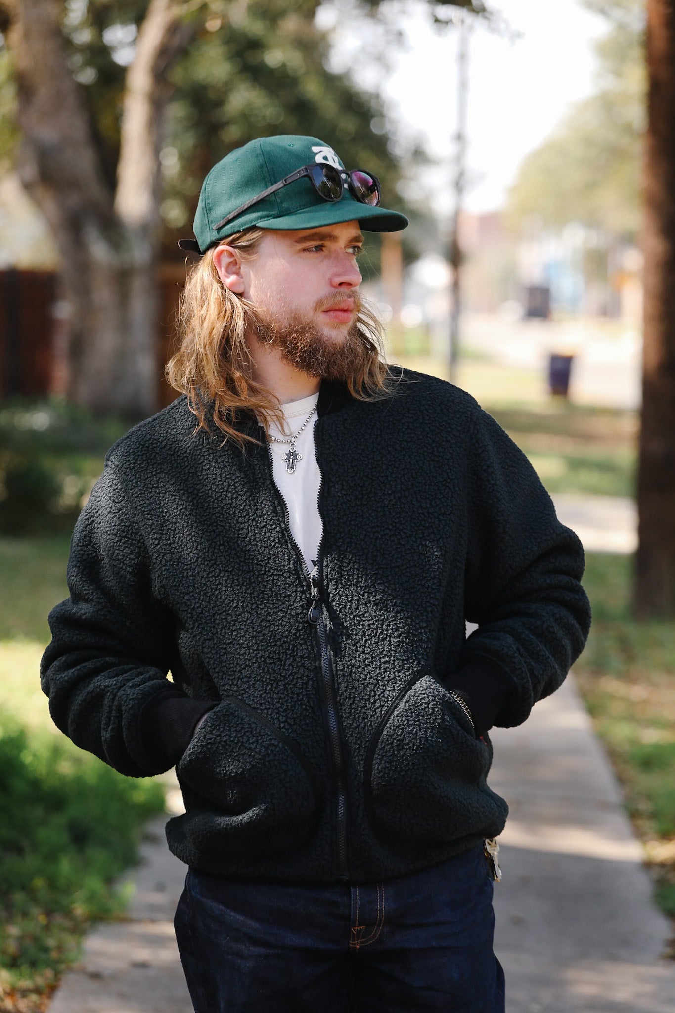 Man wearing a black fleece jacket and green cap outdoors on a path with trees and grass.