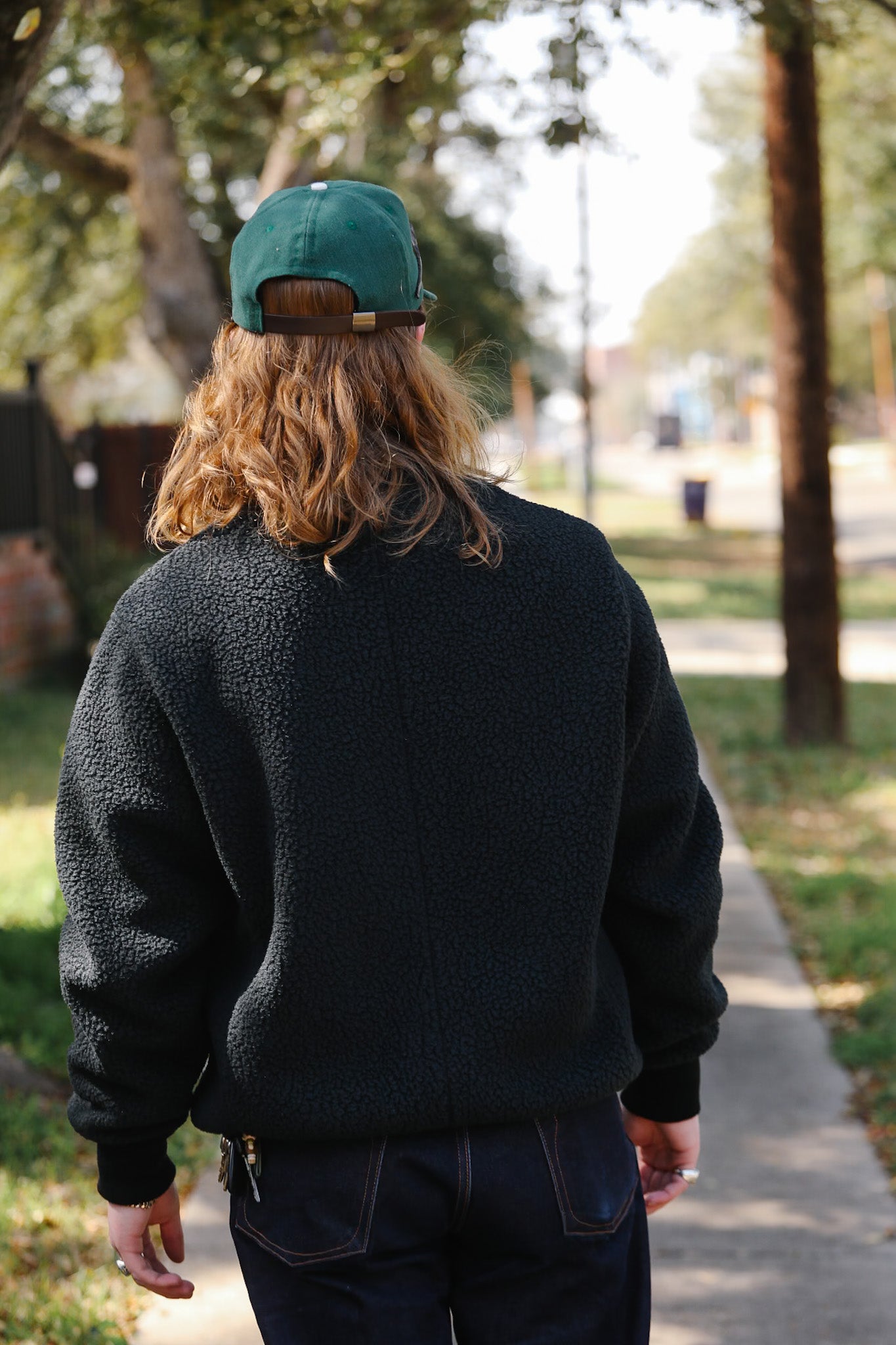 Person wearing a black jacket and teal cap walking on a path with trees and grass in the background