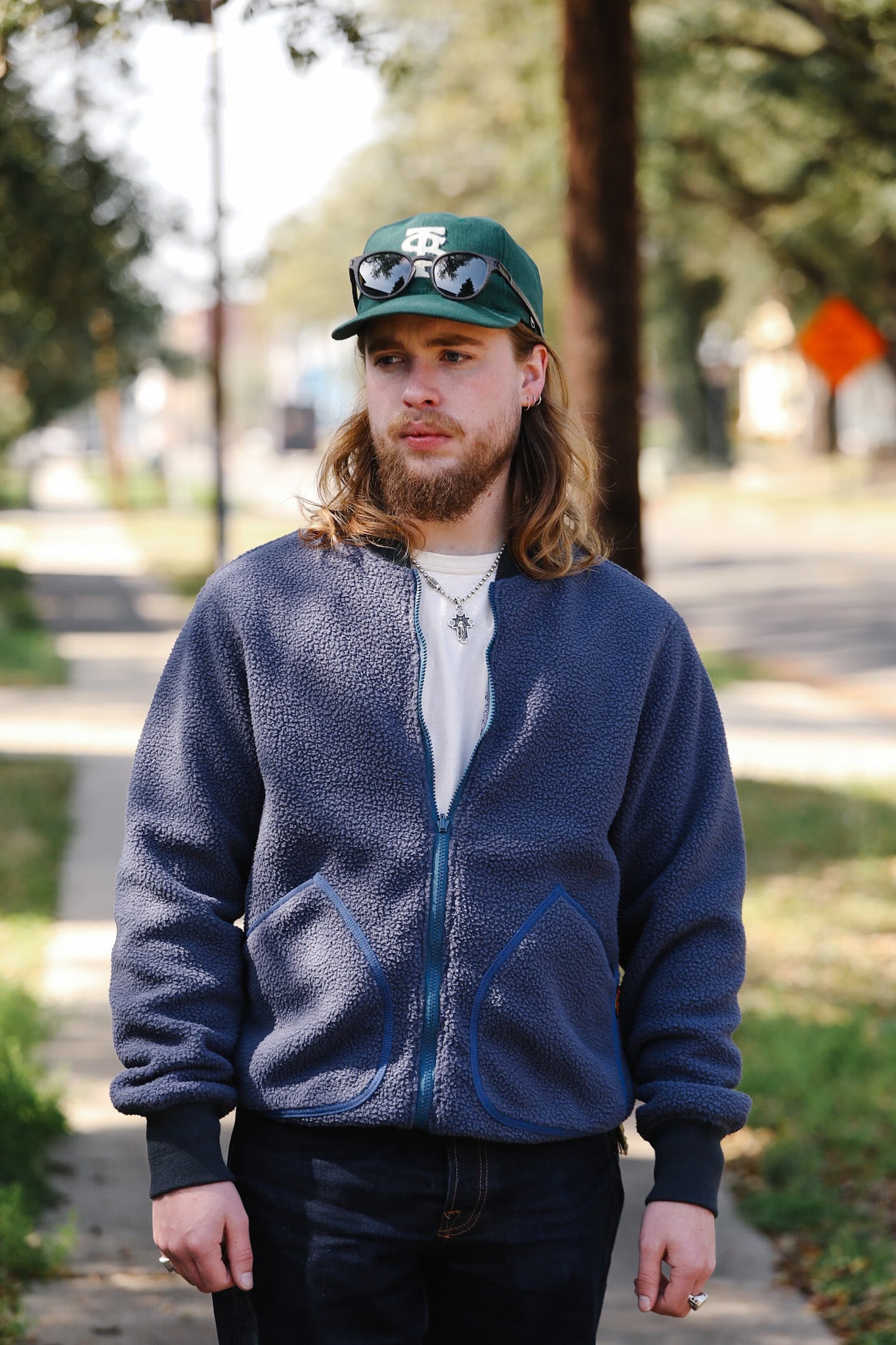 Man wearing a blue fleece jacket and green cap outdoors on a sidewalk.