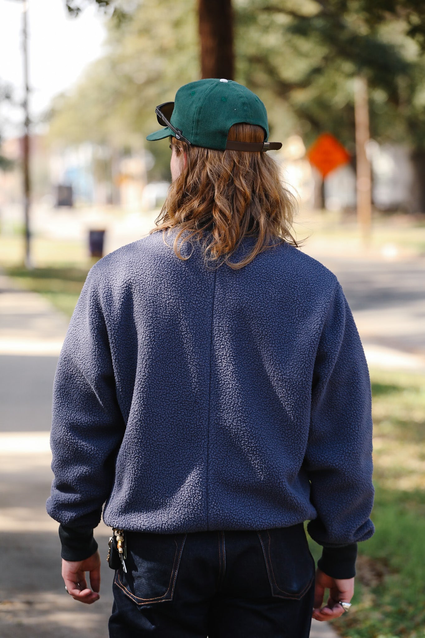 Person wearing a blue fleece jacket and green cap walking on a street with trees and a stop sign in the background.