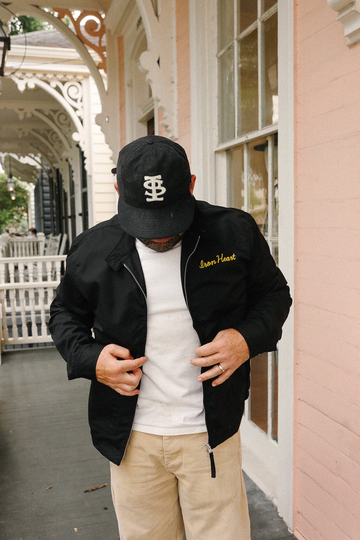 Man wearing a black jacket, white shirt, and beige pants standing in front of a pink building.