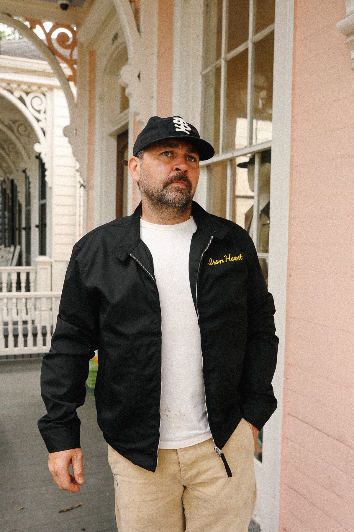 Man wearing a black jacket with yellow text, white shirt, and beige pants standing in front of a building.