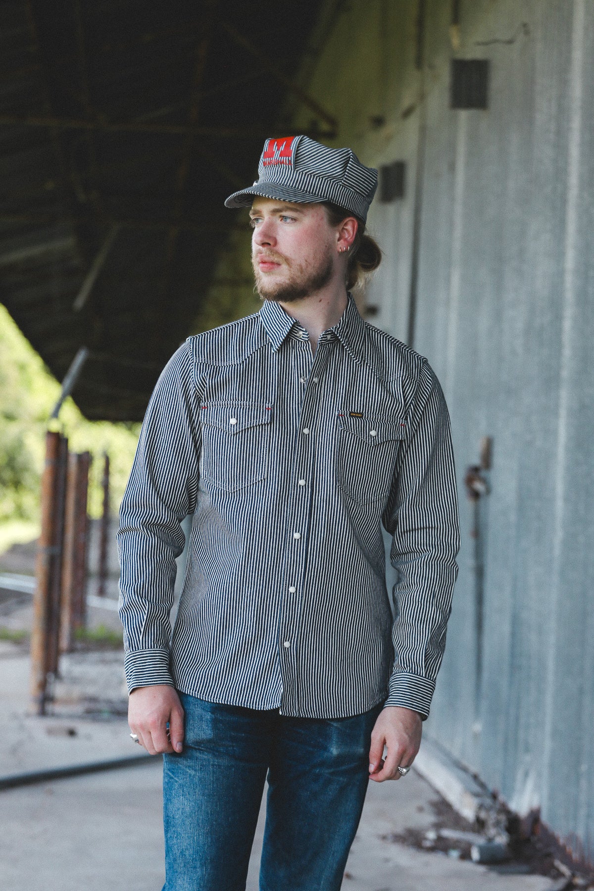 A man wearing a gray and black vertically striped long-sleeve button-down shirt, blue jeans, and a matching gray cap with a red logo patch, standing in an industrial covered walkway.