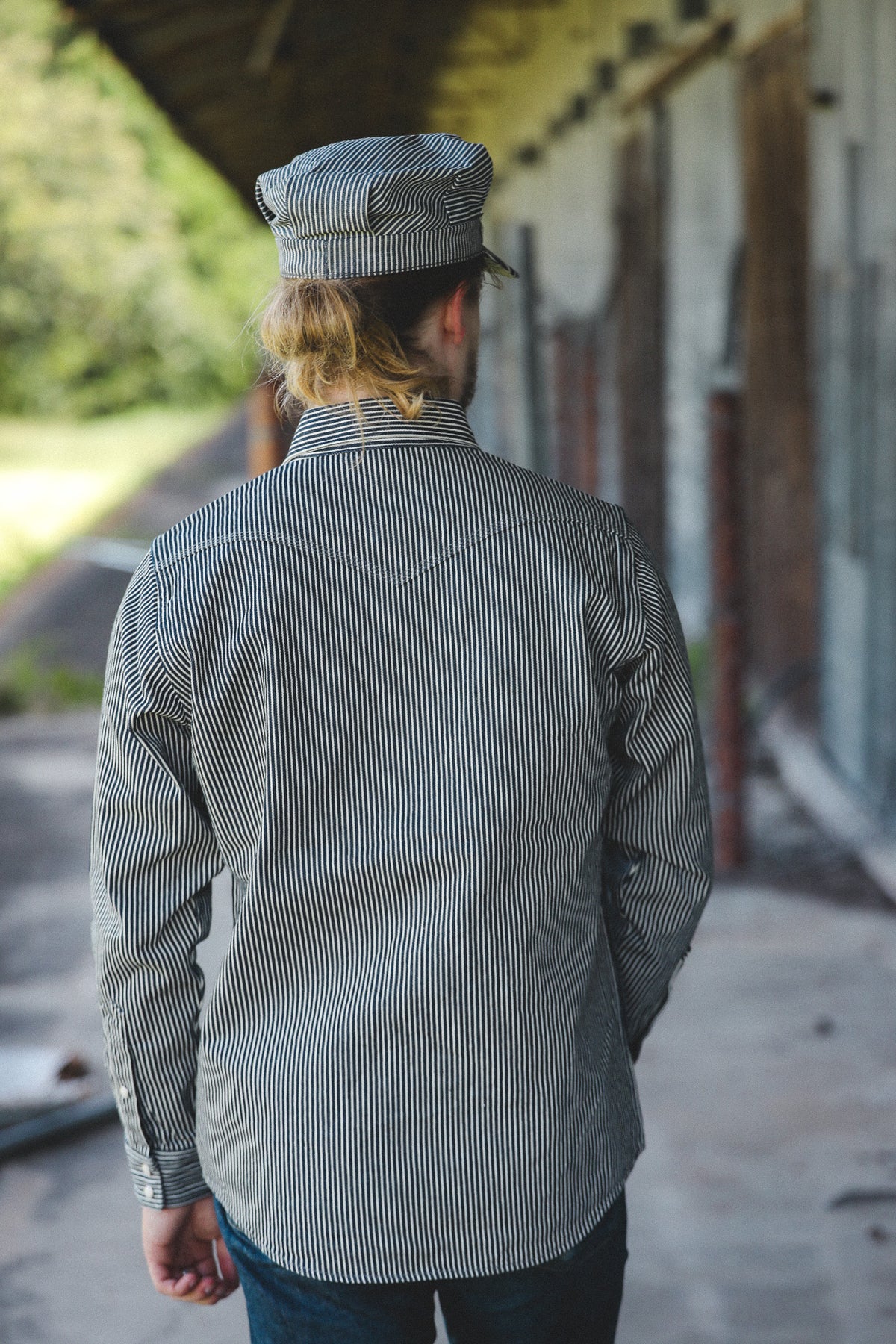 A man viewed from behind wearing a gray and white vertically striped long-sleeve shirt and matching striped cap, standing in an outdoor covered walkway.