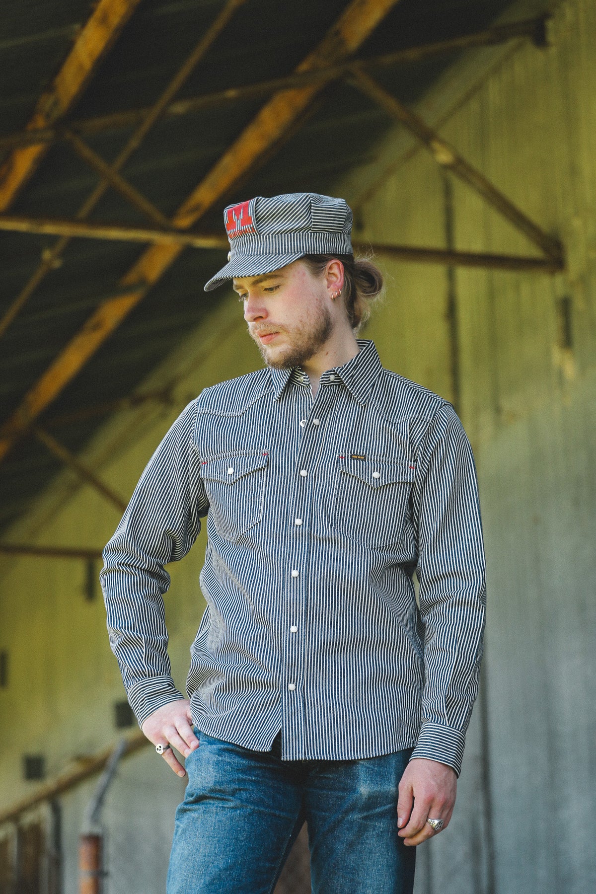 A man wearing a gray and white vertically striped long-sleeve button-down shirt, blue jeans, and a matching gray cap with a red logo, standing in a rustic barn-like environment.