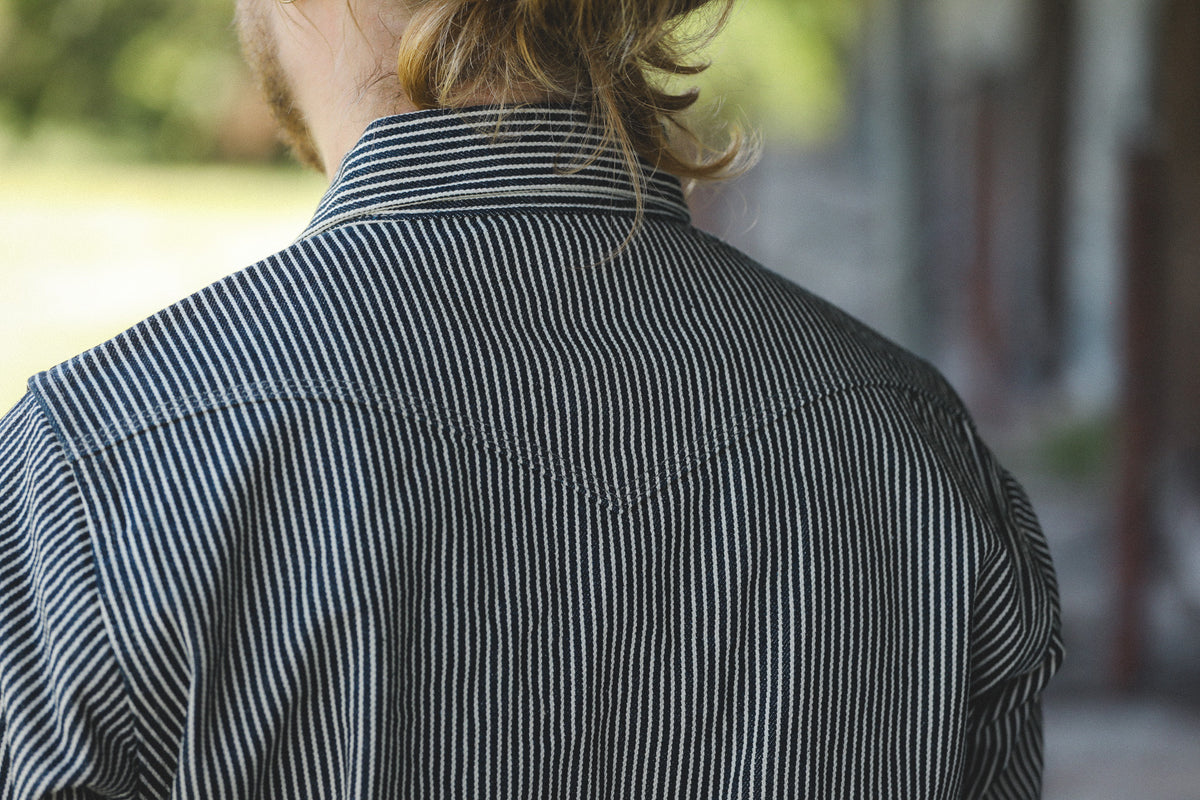 Back view of a person wearing a dark striped button-up shirt with thin vertical stripes, photographed outdoors with a blurred background.