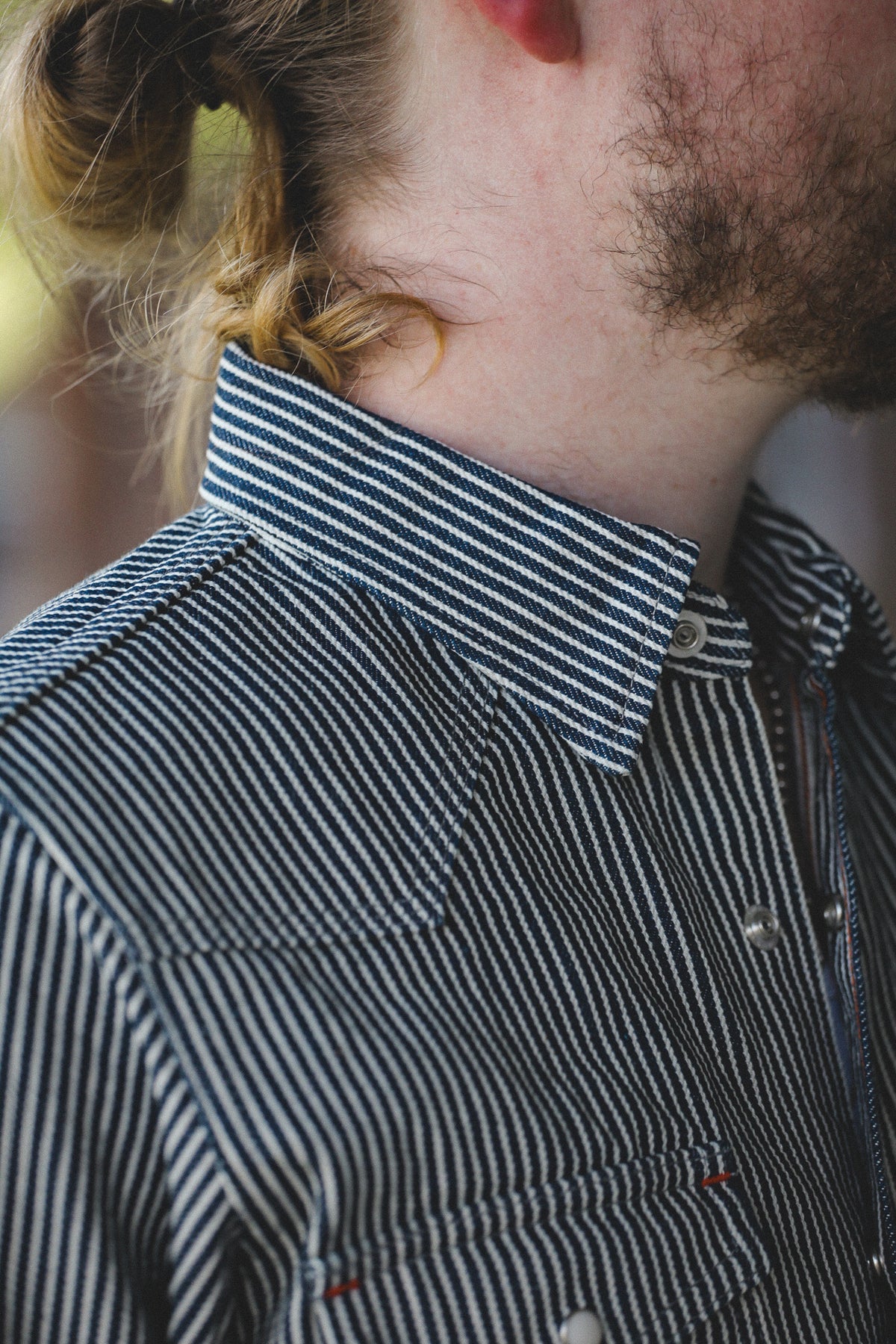 Close-up of a person wearing a dark striped button-up shirt with a pointed collar and chest pockets, showing part of the neck, beard, and tied hair.