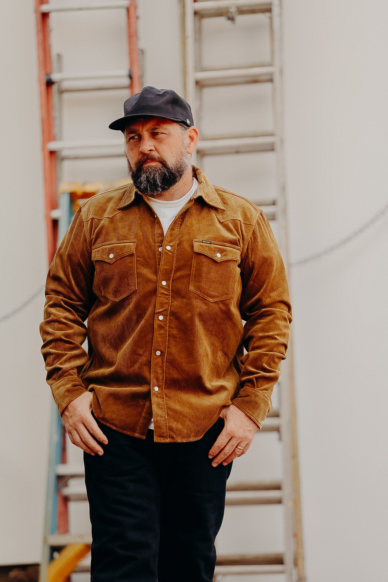 A man wearing a brown corduroy western shirt with a collar, button-up front, and cap, standing in front of a white background with a ladder in the background.