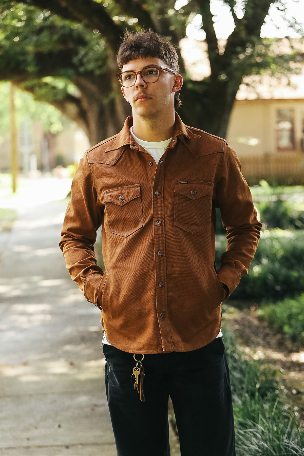 A man wearing a brown button-up shirt jacket over a white t-shirt and black pants, standing on a tree-lined path.