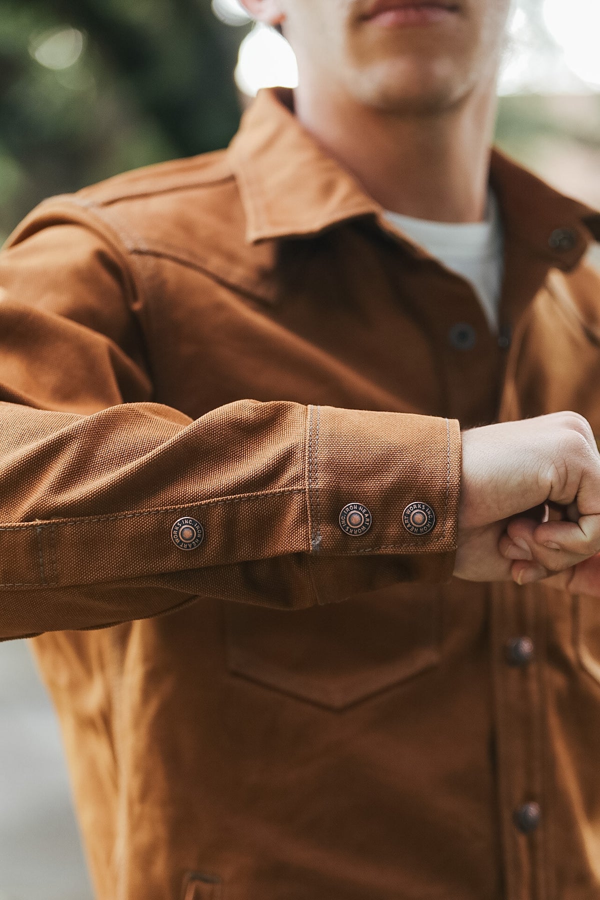 A close-up of a person wearing a brown suede or nubuck utility jacket over a white t-shirt, with the person's hands adjusting the cuff of the jacket.