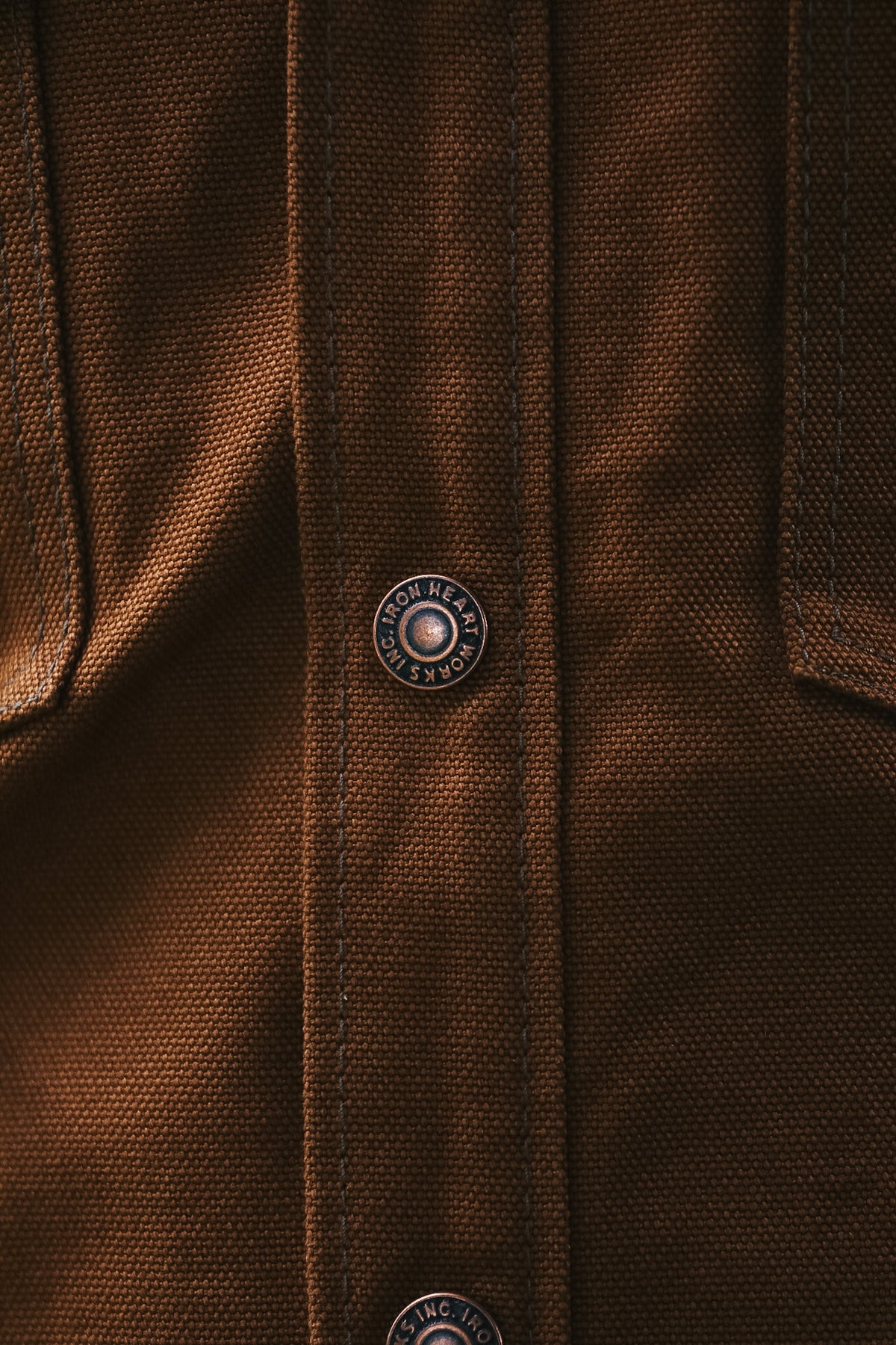 Close-up of a brown textured fabric garment with a vertical button placket and a visible round metal button engraved with the brand name 'HARROSH'.