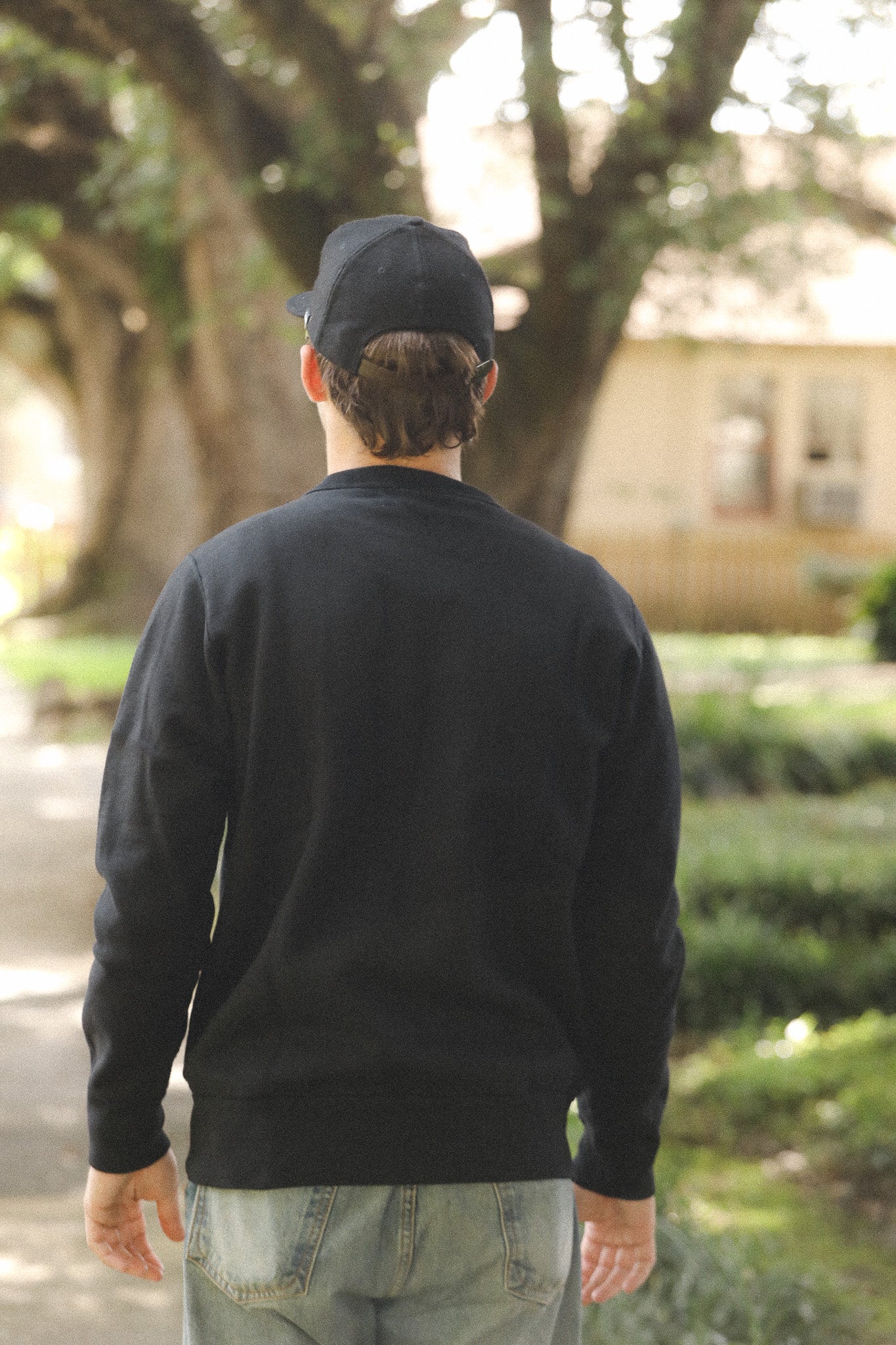 Person wearing a black sweatshirt and cap walking outdoors with trees and a building in the background