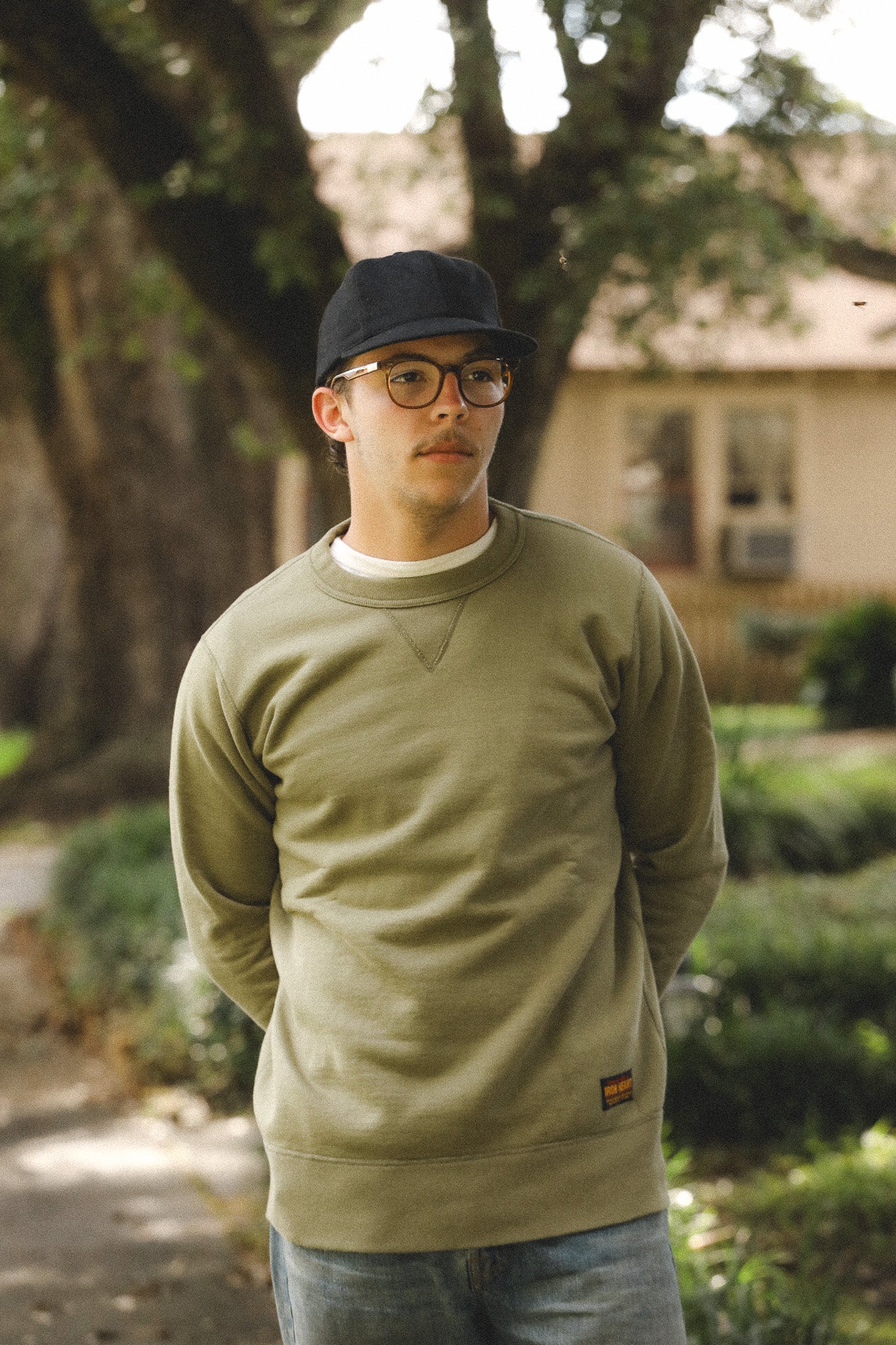 Man wearing a green sweatshirt and cap standing outdoors with trees and a house in the background