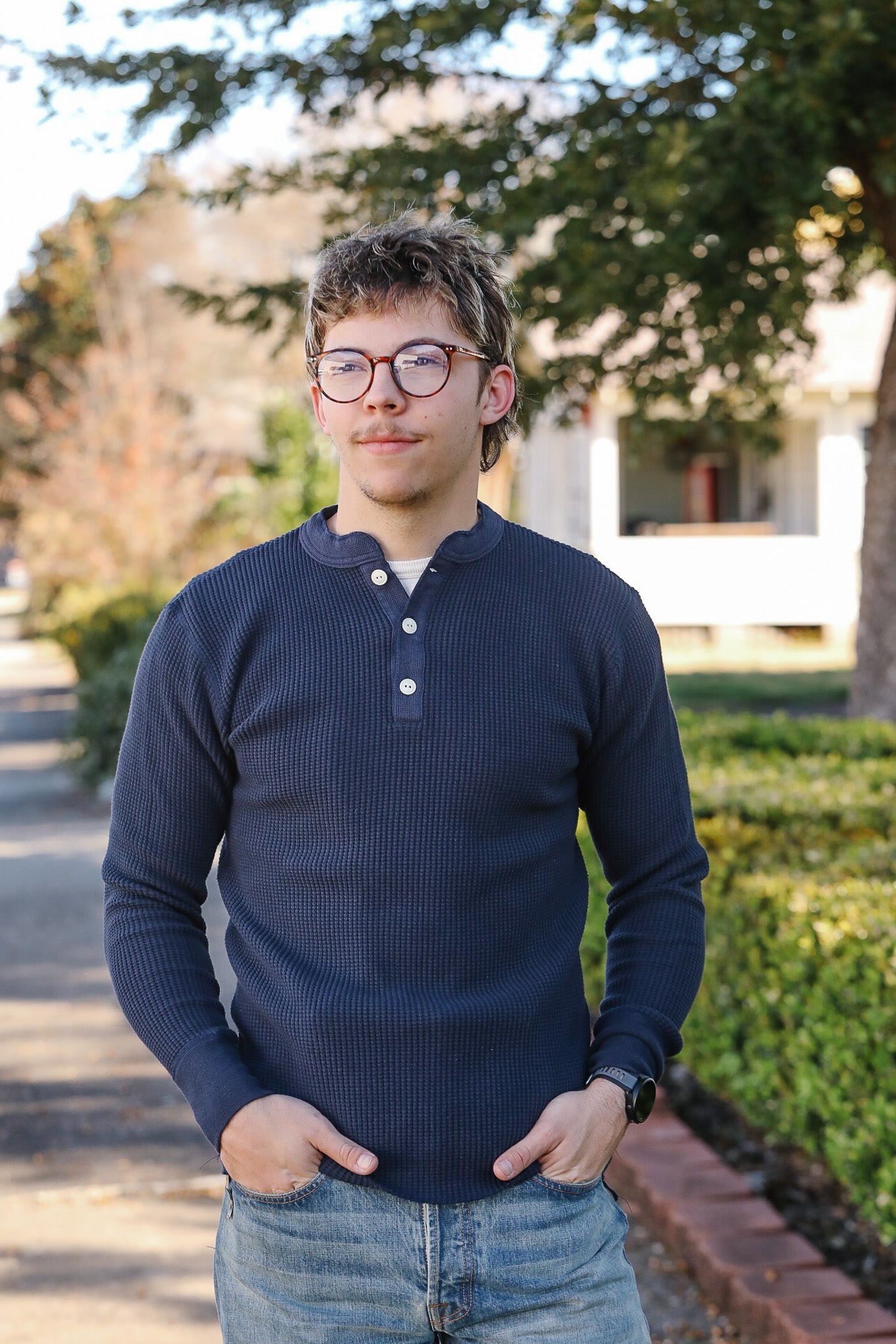 Person wearing a navy blue long-sleeve shirt standing outdoors with trees and a building in the background.