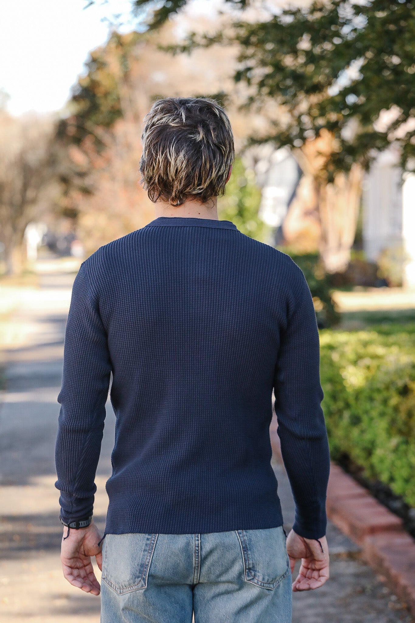 Man wearing a navy sweater and jeans walking away from the camera in a park-like setting.