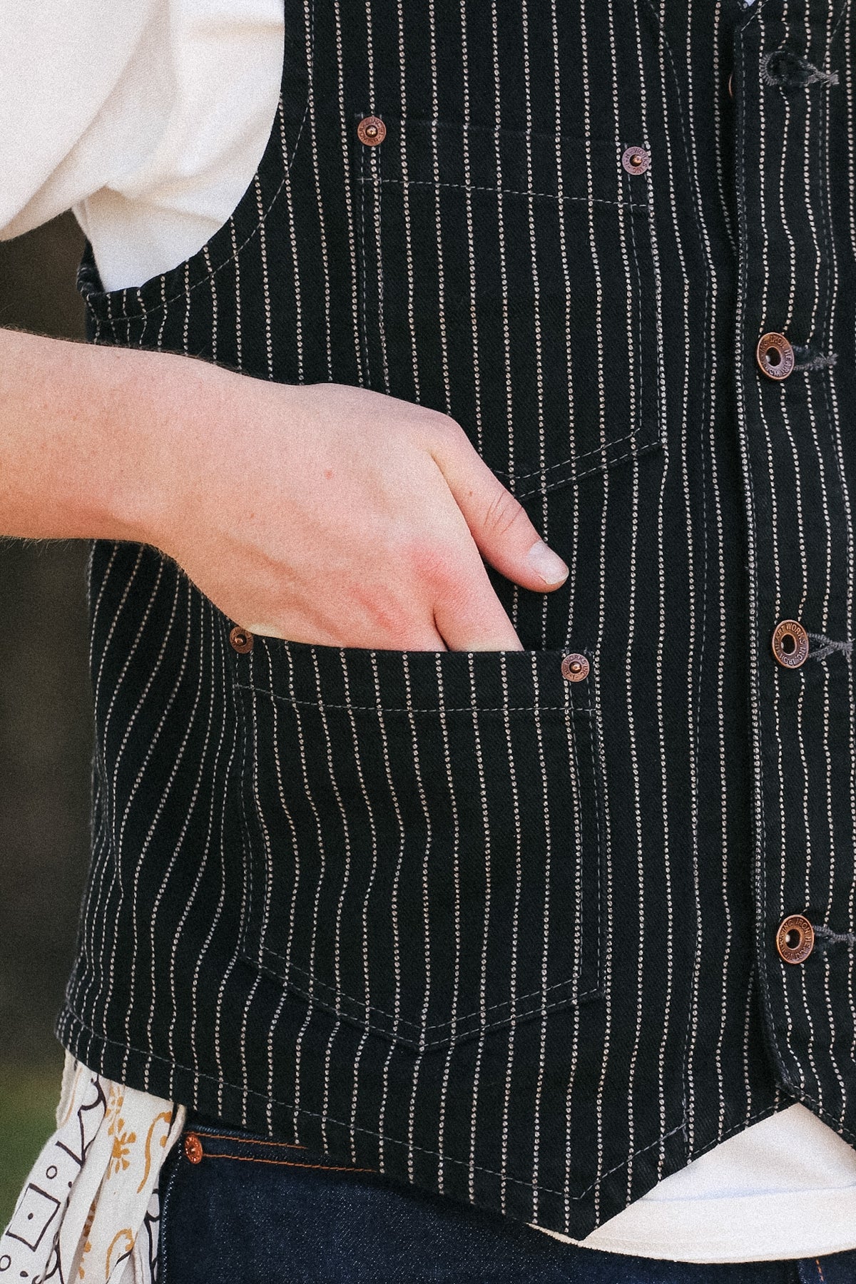 Close-up of a person wearing a black vest with thin white vertical pinstripes, brown buttons, and a front patch pocket, with a hand reaching into the pocket.
