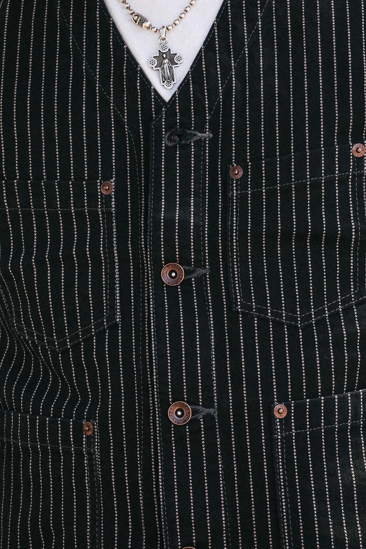 Close-up of a black pinstripe vest with copper-colored buttons, worn over a white shirt and a silver cross pendant necklace.