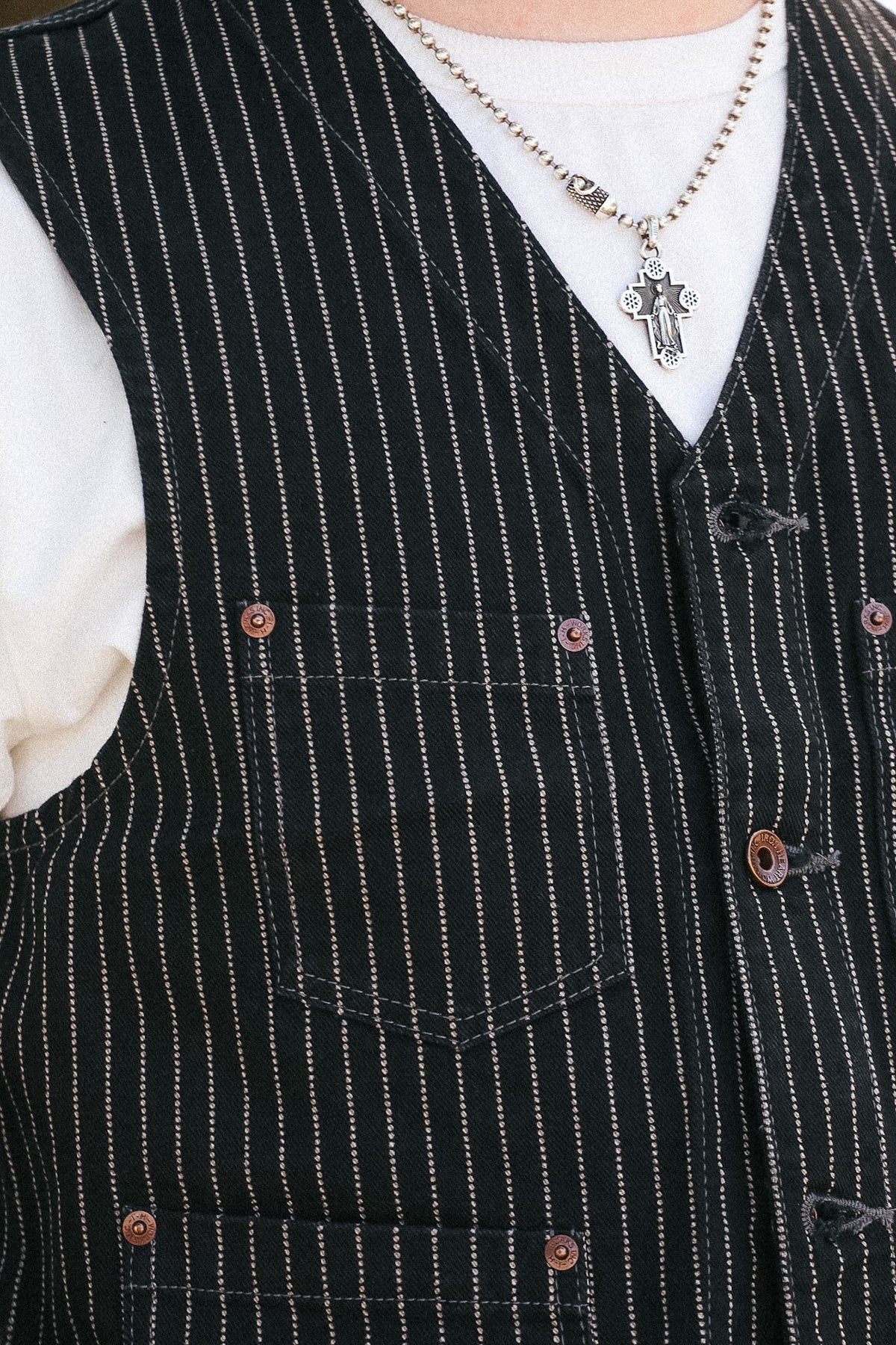 Close-up of a black pinstripe vest with brown buttons, worn over a white t-shirt and a silver cross necklace.