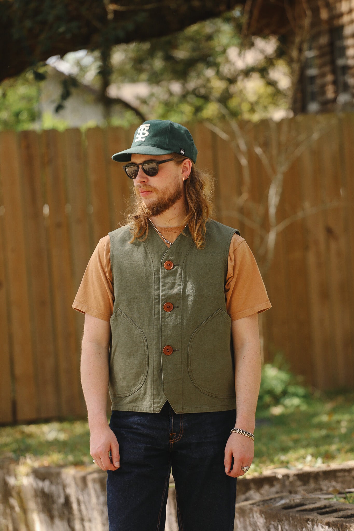 Man wearing a green vest and cap outdoors with a wooden fence and trees in the background