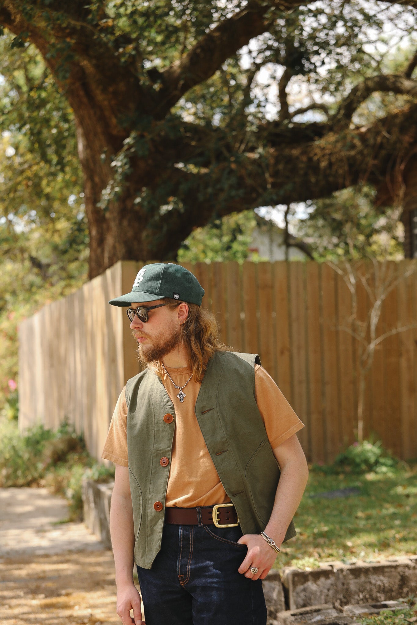 Man wearing a green cap, vest, and sunglasses outdoors with trees and a wooden fence in the background.