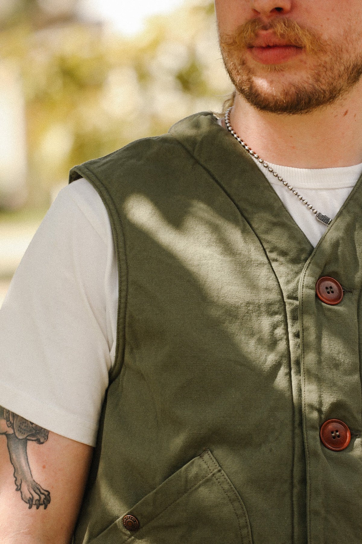 A close-up of a man wearing an olive green utility vest over a white t-shirt, with a tattoo visible on his arm and a blurred outdoor background.