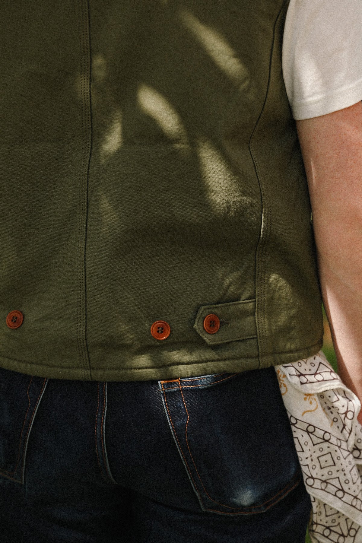 Close-up of a person wearing an olive green sleeveless jacket or vest with brown buttons, paired with dark blue jeans and a white sleeveless shirt. Sunlight casts shadows across the fabric.