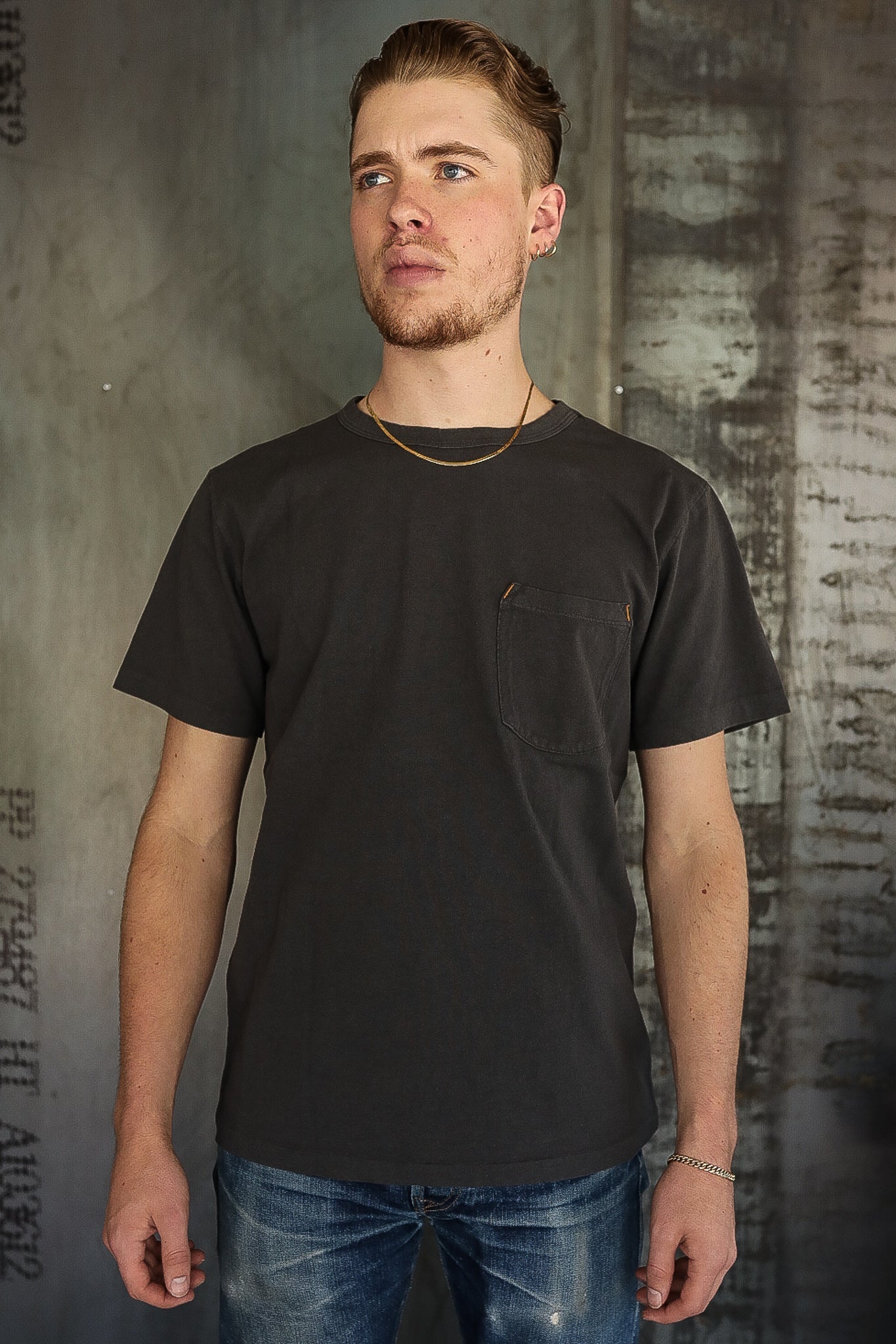 Young man wearing black pocket t-shirt and blue jeans standing against textured industrial wall at Iron Shop Provisions