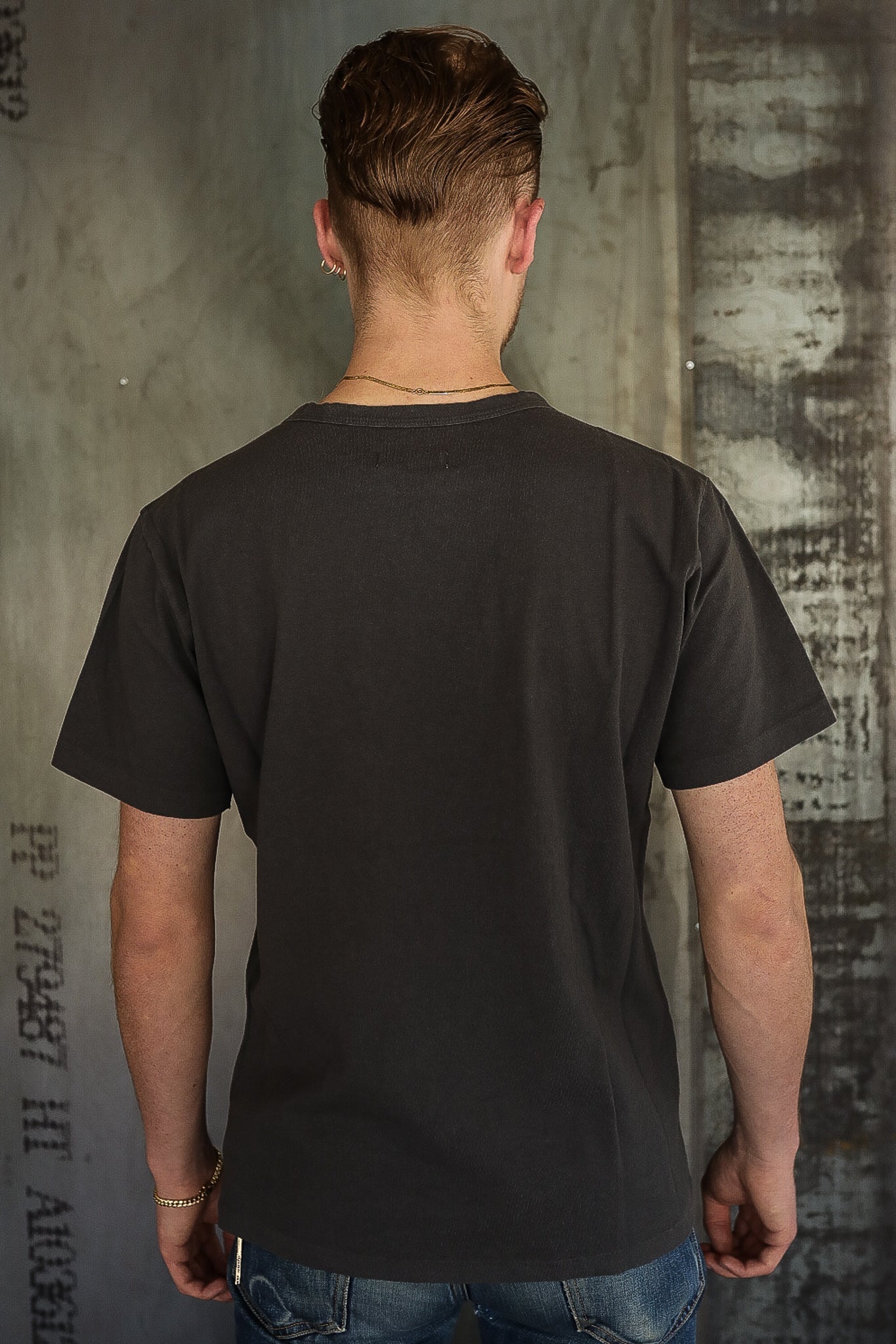 Man wearing plain black short-sleeve t-shirt and blue jeans standing against industrial concrete wall