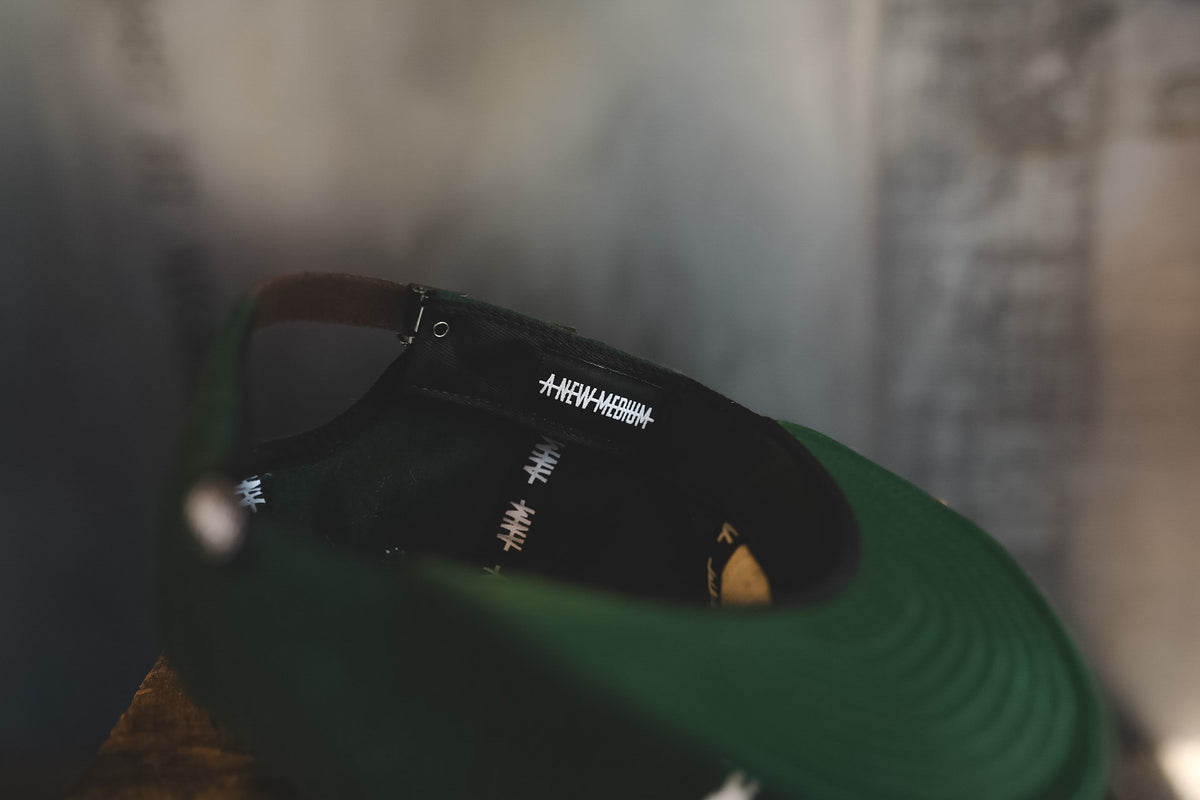 A close-up shot of a dark green baseball cap with a black interior, featuring white embroidered text on the inside and a silver metal snap on the brim.