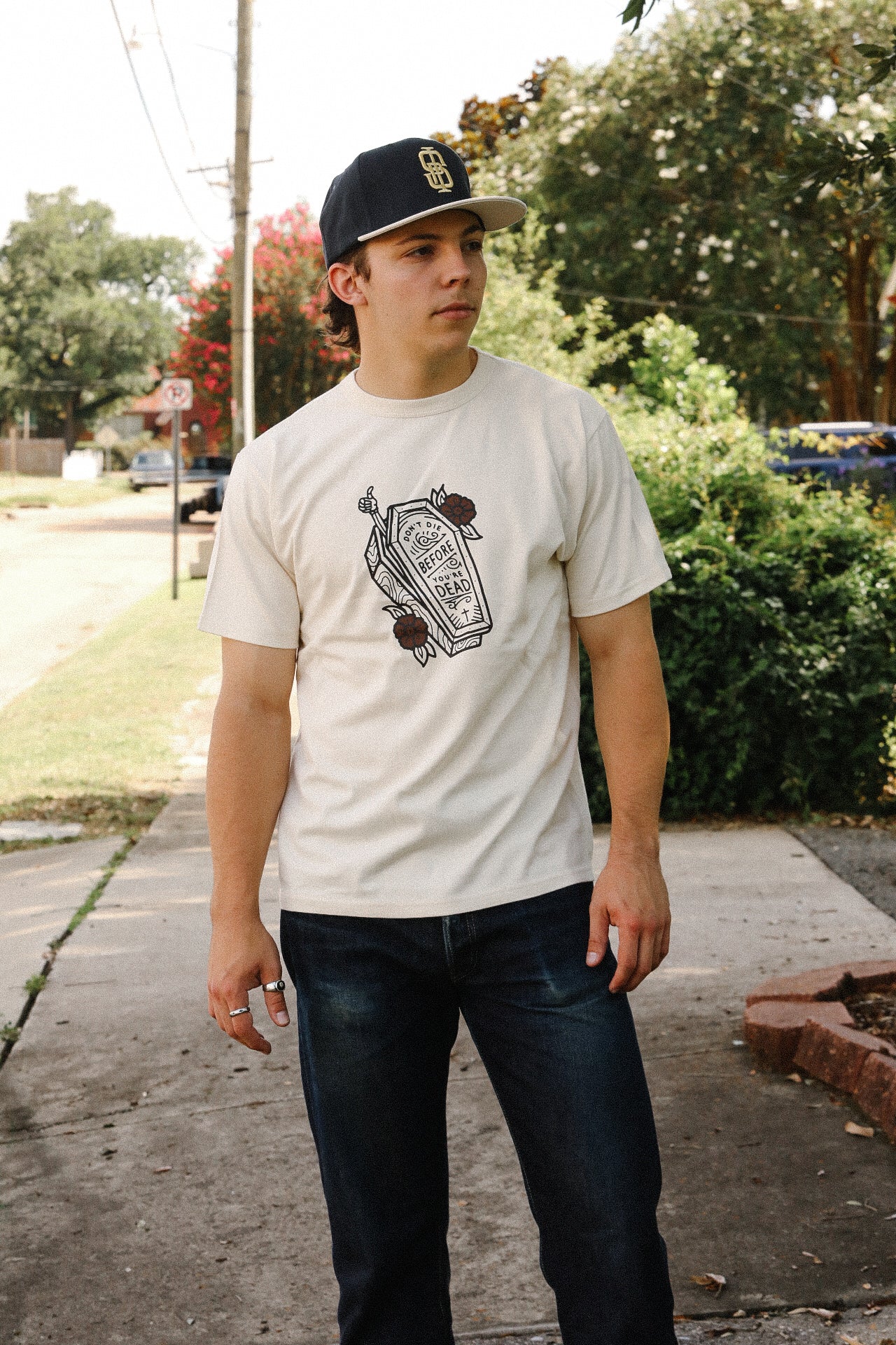 Young man wearing cream graphic t-shirt with coffin design and black baseball cap, standing on sidewalk with greenery background