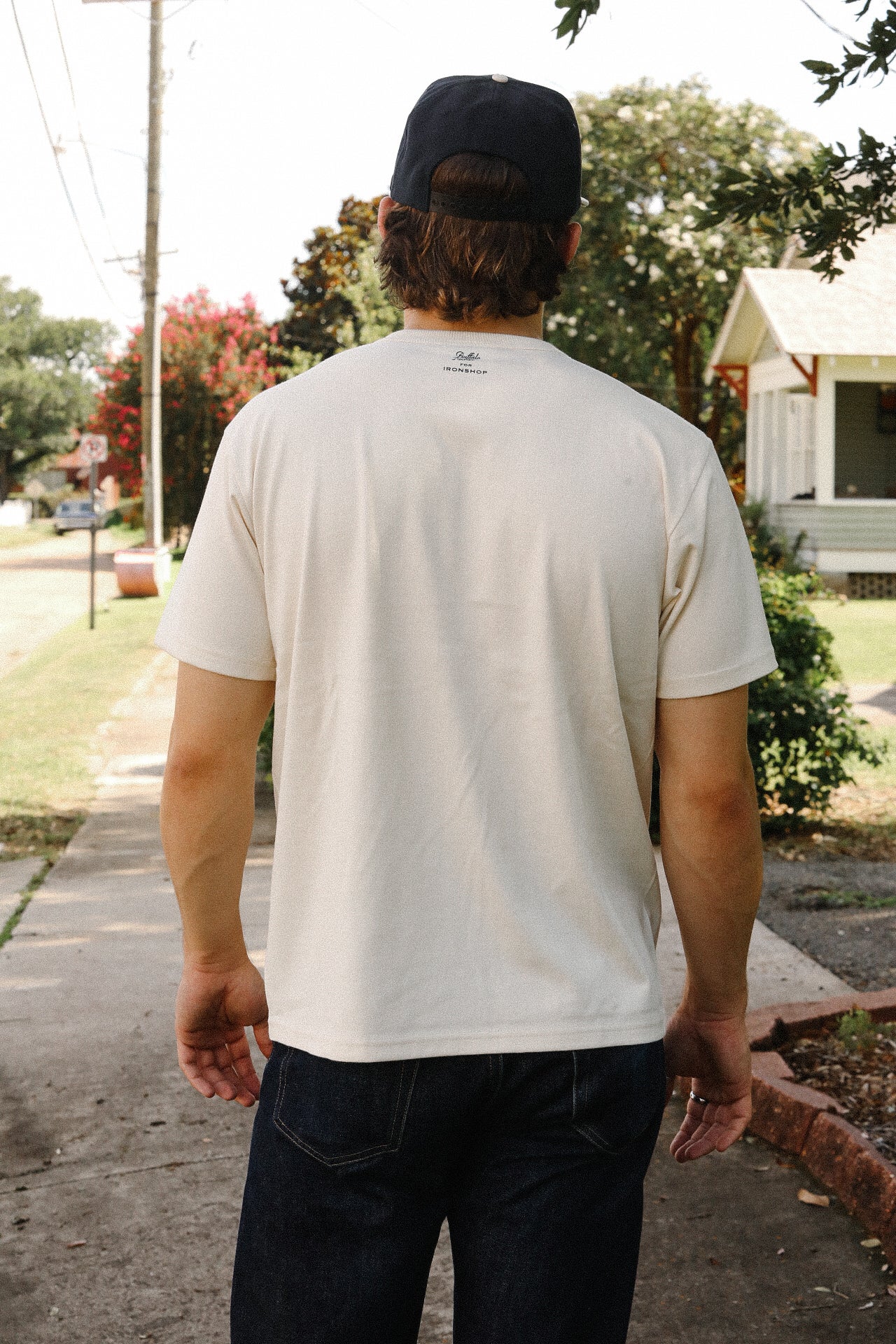 Man wearing cream Iron Shop Provisions t-shirt and dark jeans walking on suburban sidewalk with trees and houses