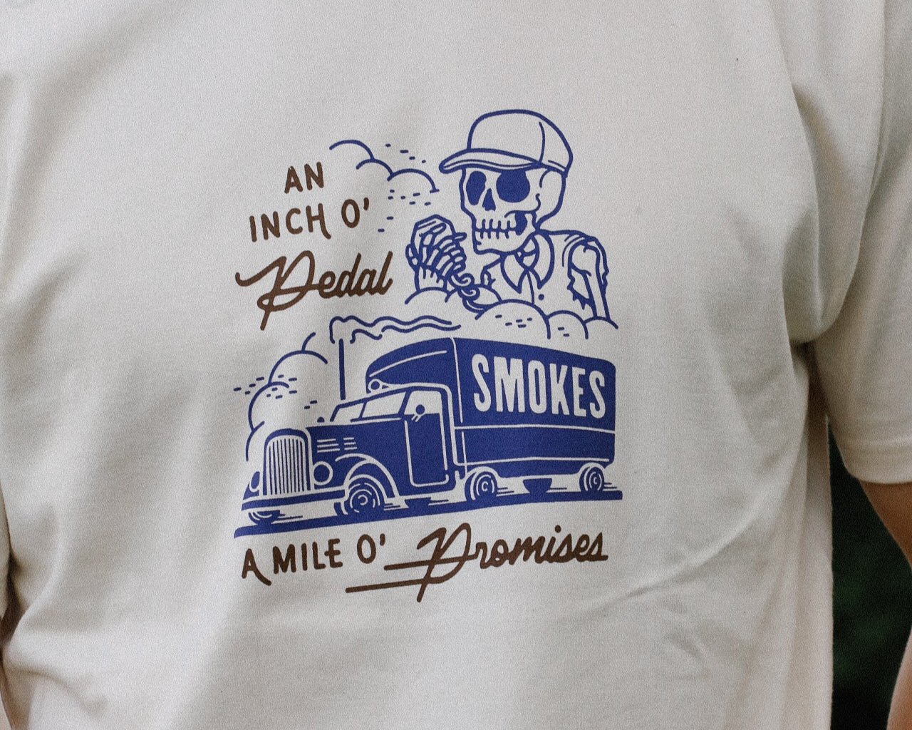 Cream-colored Iron Shop Provisions t-shirt featuring blue graphic of a skeleton driver and truck with smoke and text reading an inch o' pedal a mile o' promises