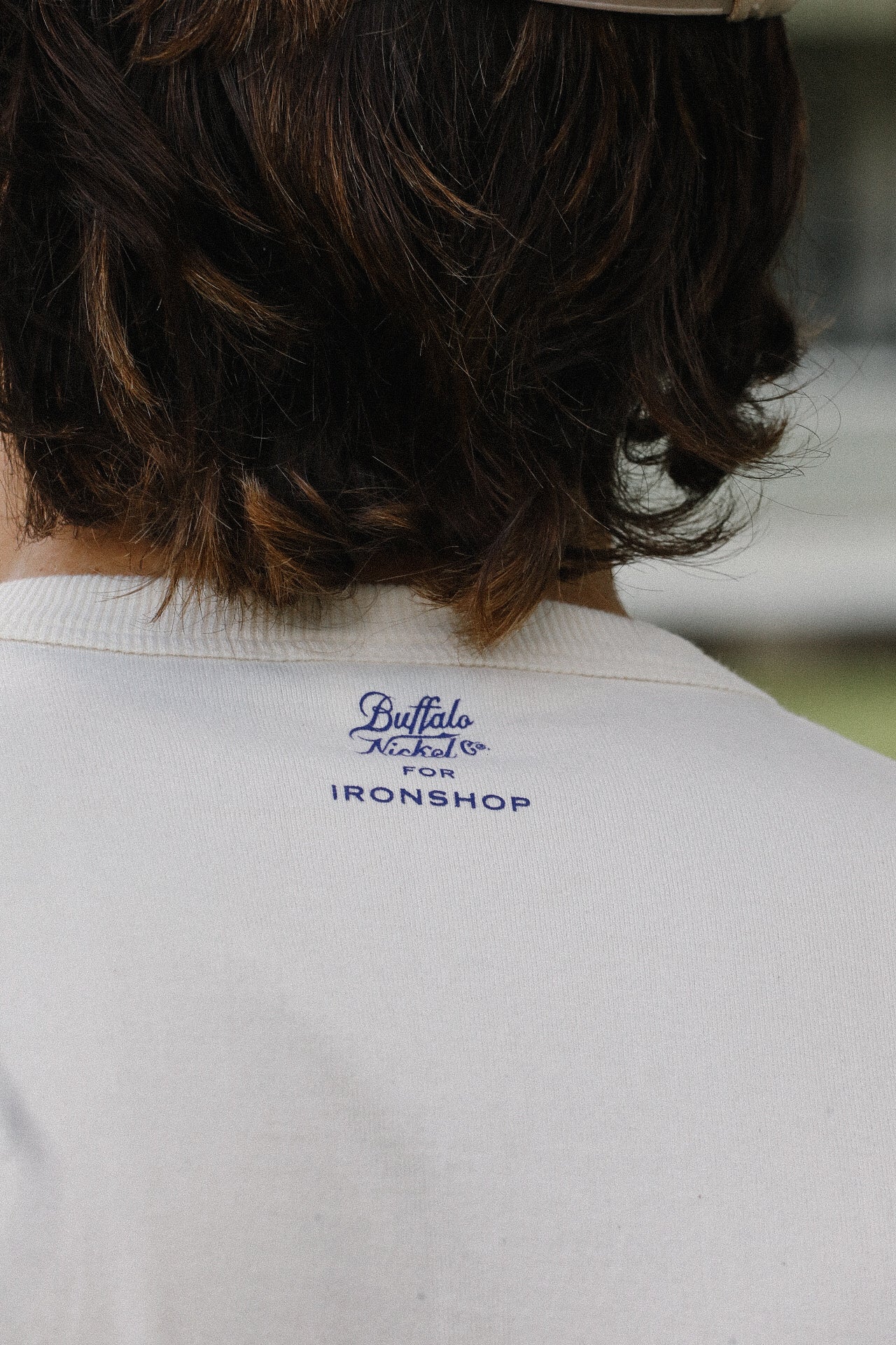 Close-up of cream loopwheel tee back with Buffalo Nickel Co logo for Ironshop under brown wavy hair