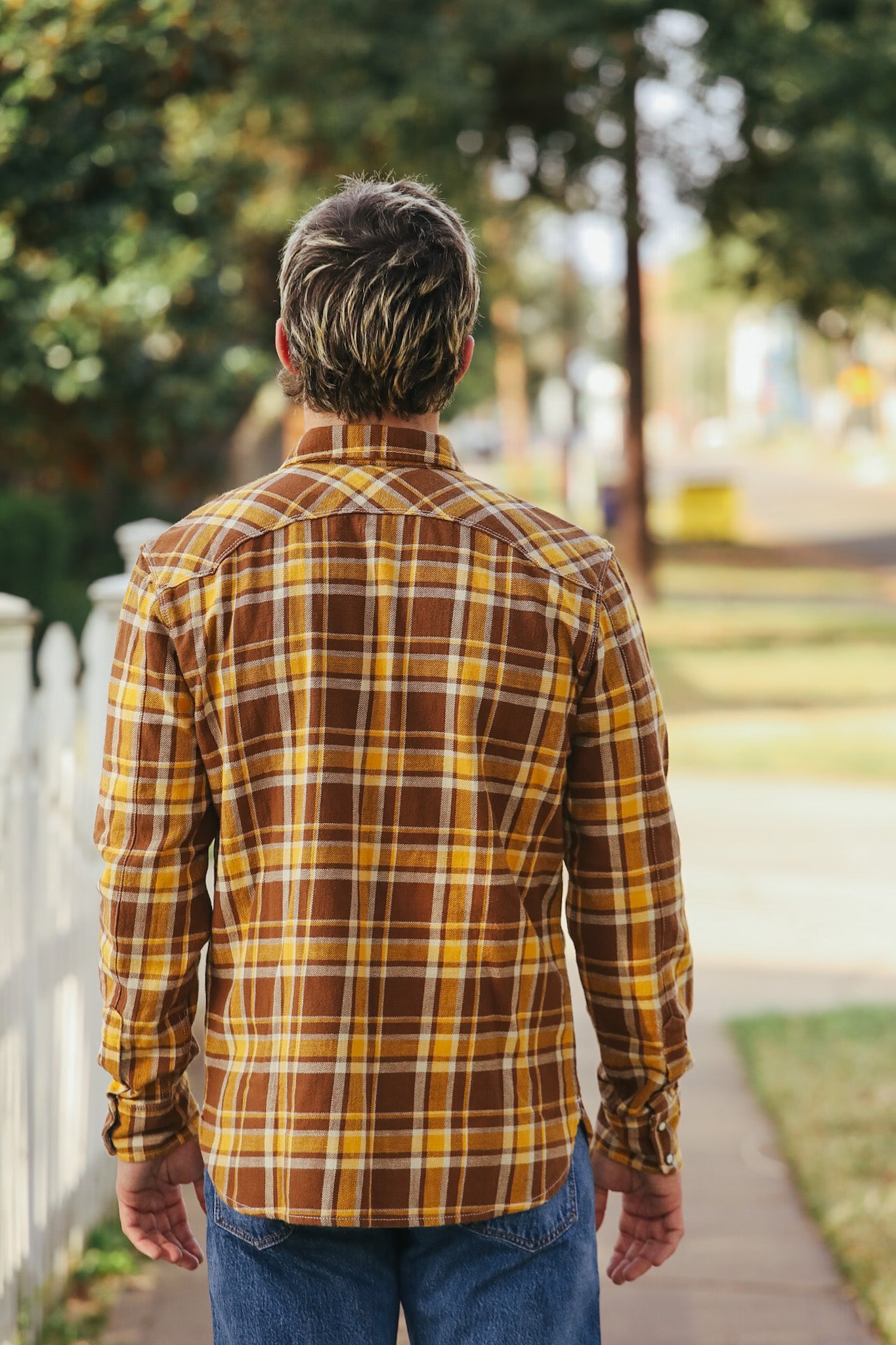 Man wearing a plaid shirt walking away on a path with trees and grass in the background
