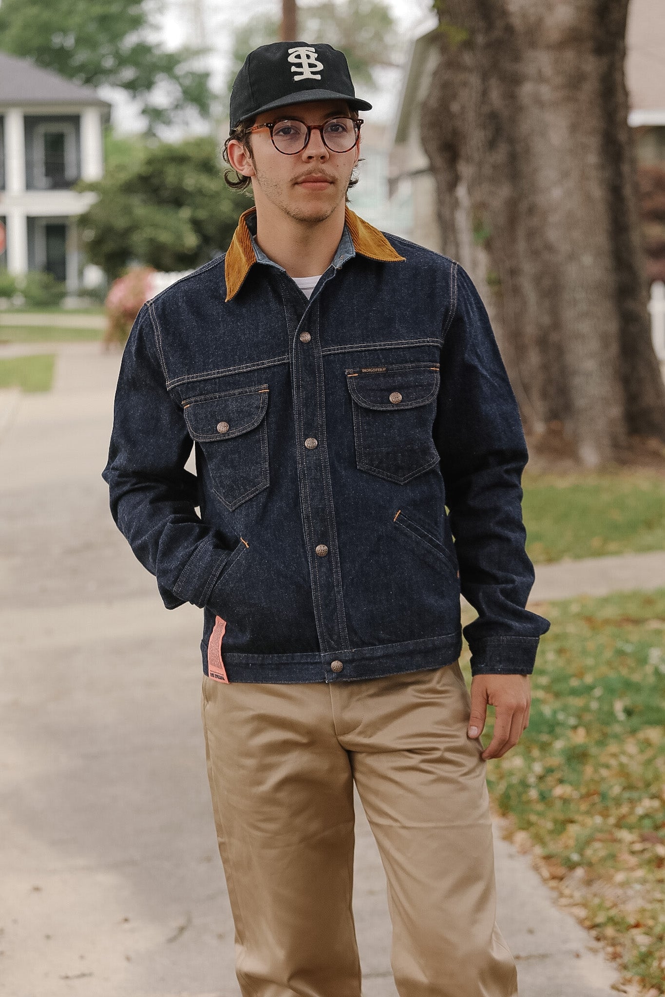 Man wearing a denim jacket and beige pants standing on a sidewalk.