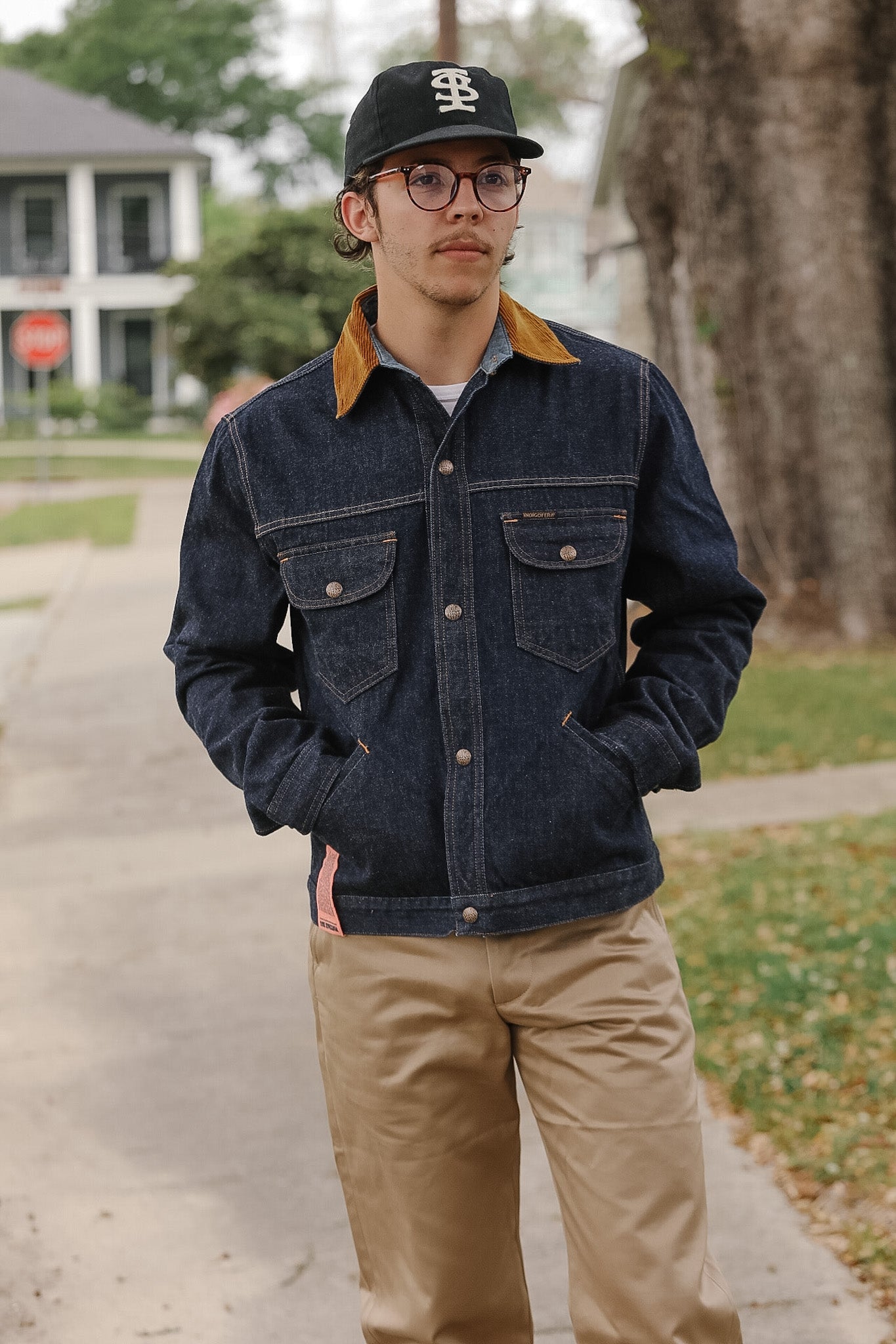 Person wearing a denim jacket and cap standing on a sidewalk with trees and houses in the background