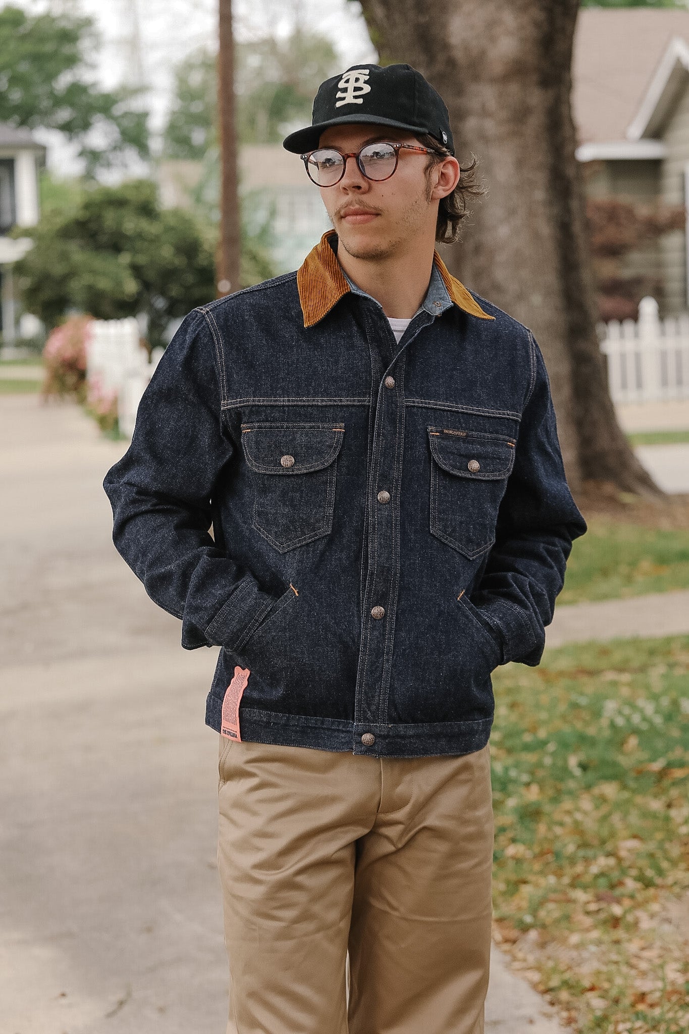 Man wearing a denim jacket, cap, and glasses standing outdoors.