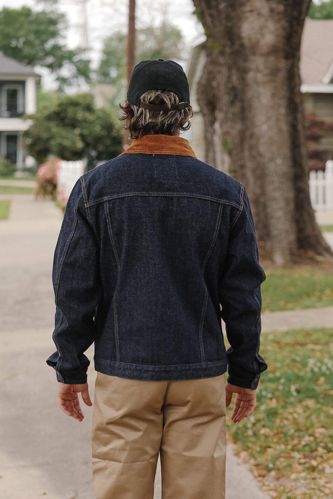 Person wearing a dark denim jacket and beige pants walking on a sidewalk with trees and houses in the background.