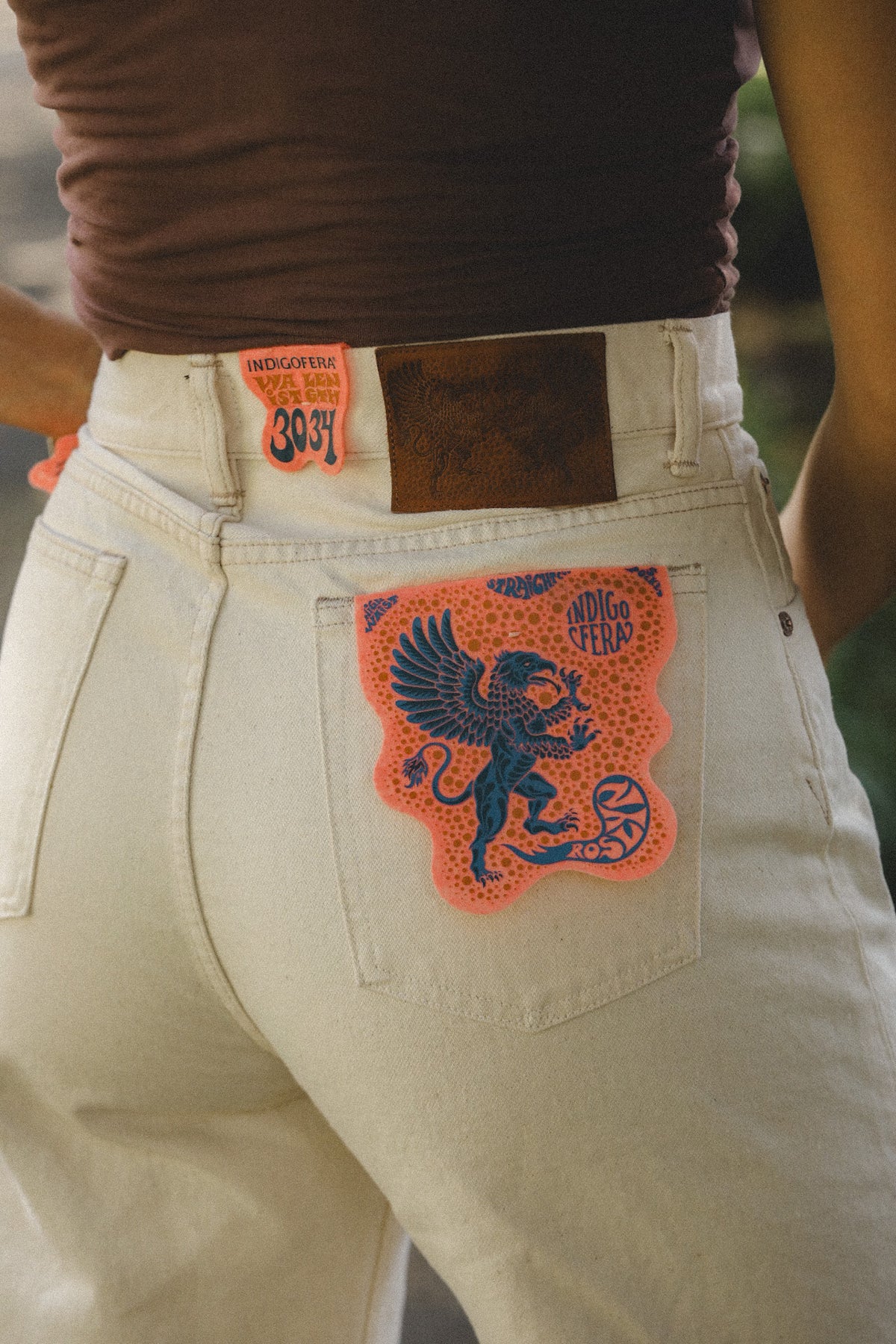 Close-up of the back of a person wearing off-white denim jeans with a large orange and blue graphic patch on the back pocket, plus a brown leather waistband patch.