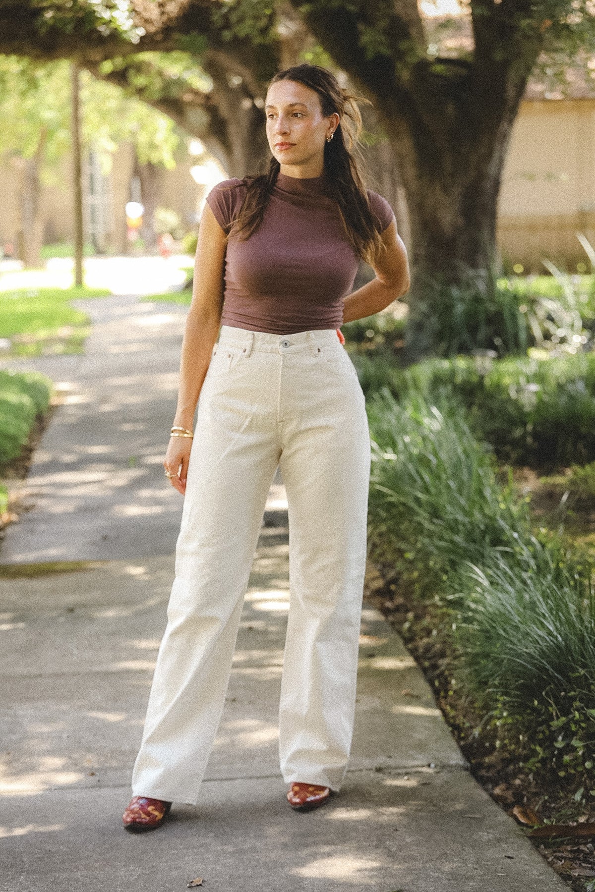 A woman stands on a tree-lined sidewalk wearing a fitted short-sleeve mauve-brown mock-neck top and high-waisted off-white wide-leg jeans, paired with brown pointed flats.